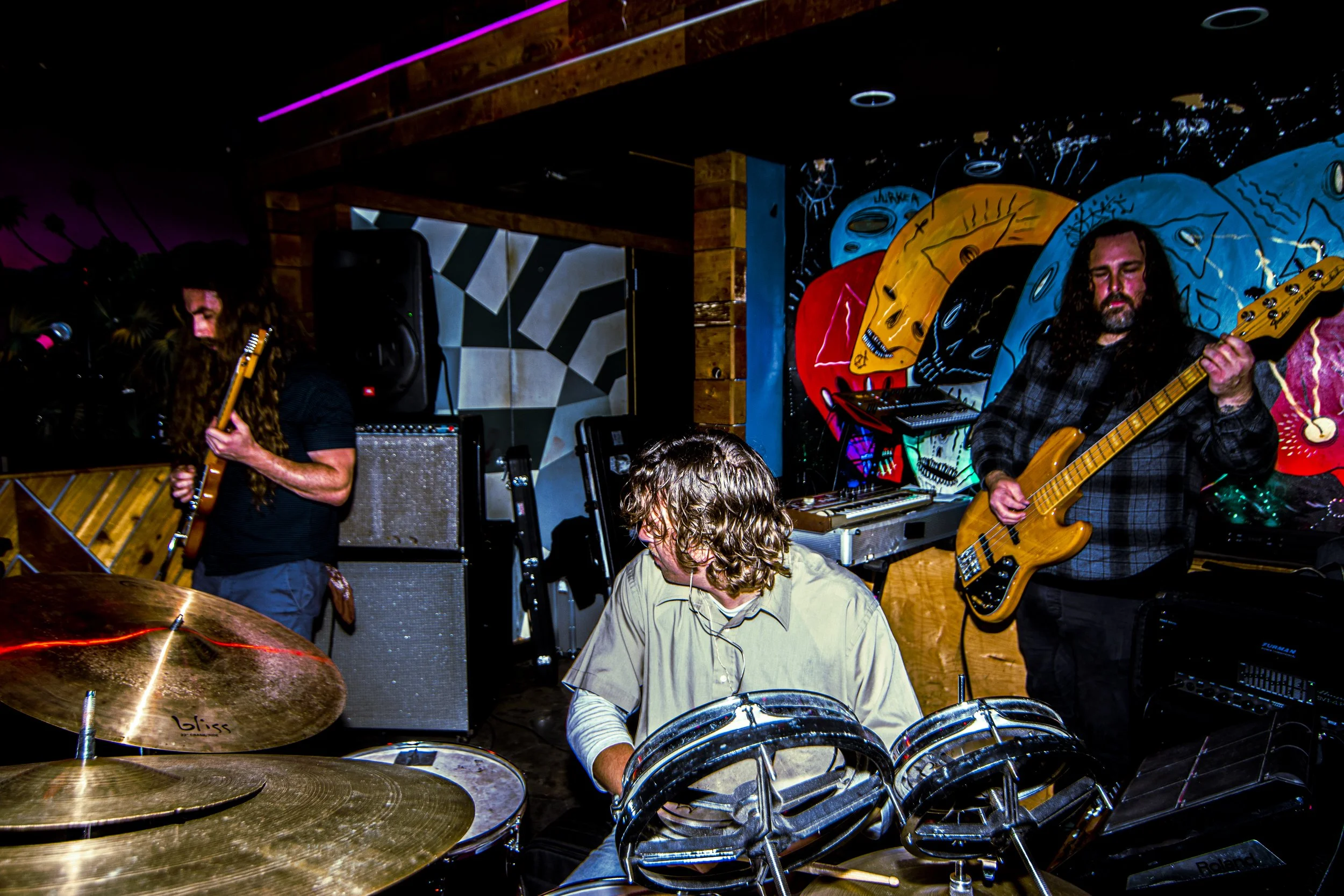 Three musicians performing: a guitarist on the left, a drummer in the center, and a bassist on the right, in a colorful, artistic venue with vibrant wall art.