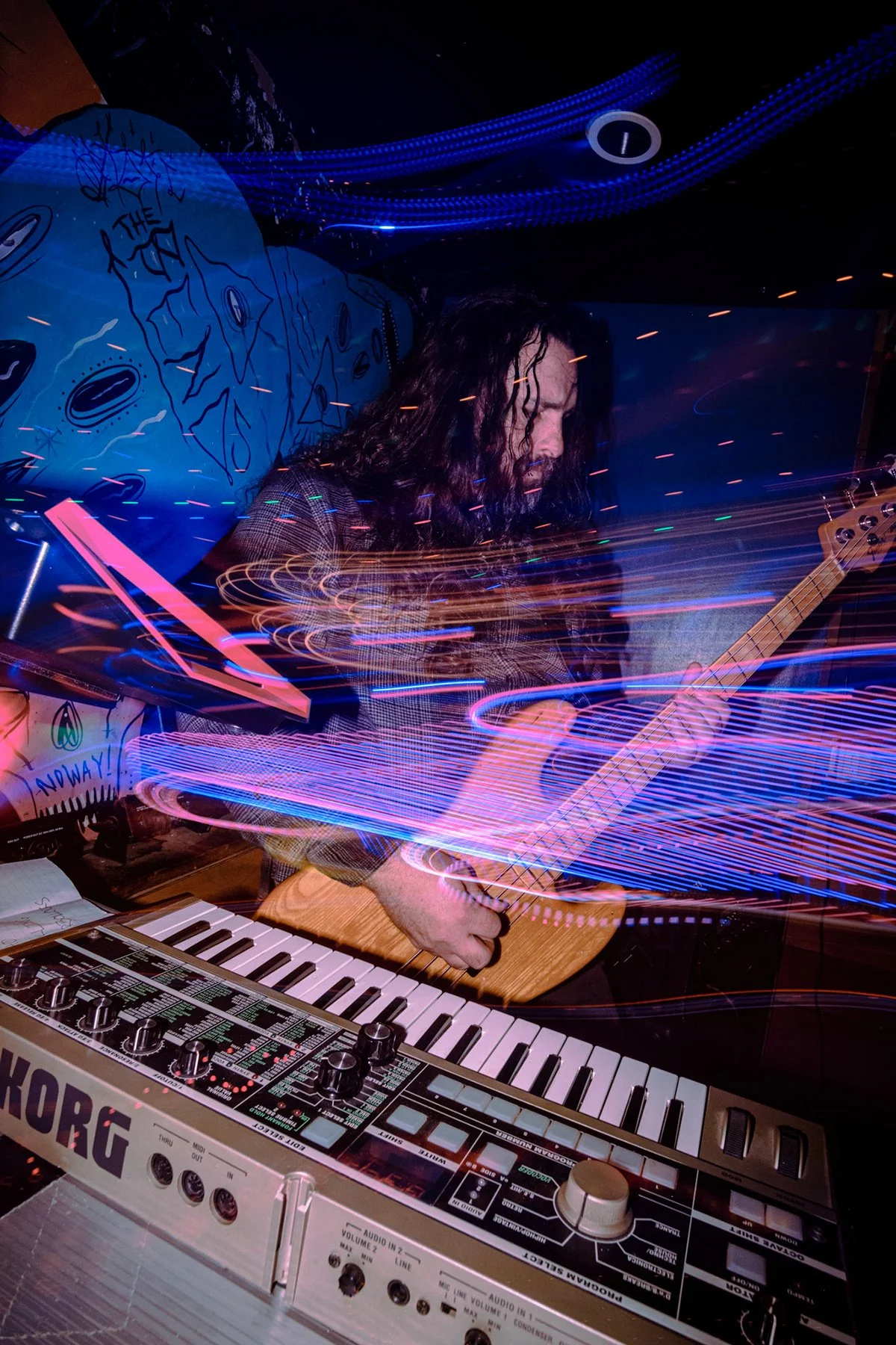 Tysonious Mink of Liquid Pennies plays bass and keyboard with colorful light trails and abstract wall art in the background.