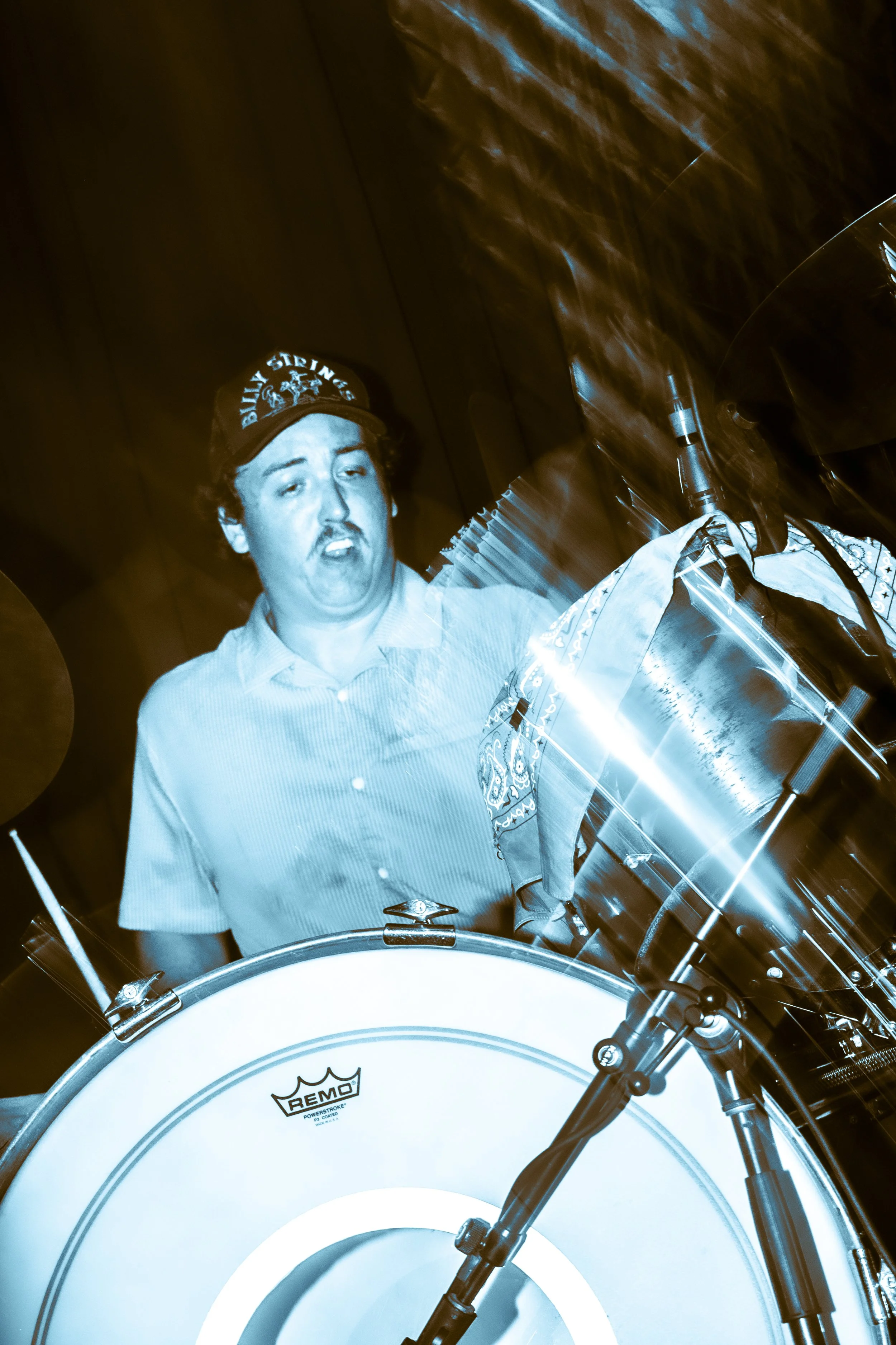 A person playing a drum set with a distressed facial expression, wearing a cap with the words 'Billy Strings' on it. The image has a blue tint and motion blur effects.