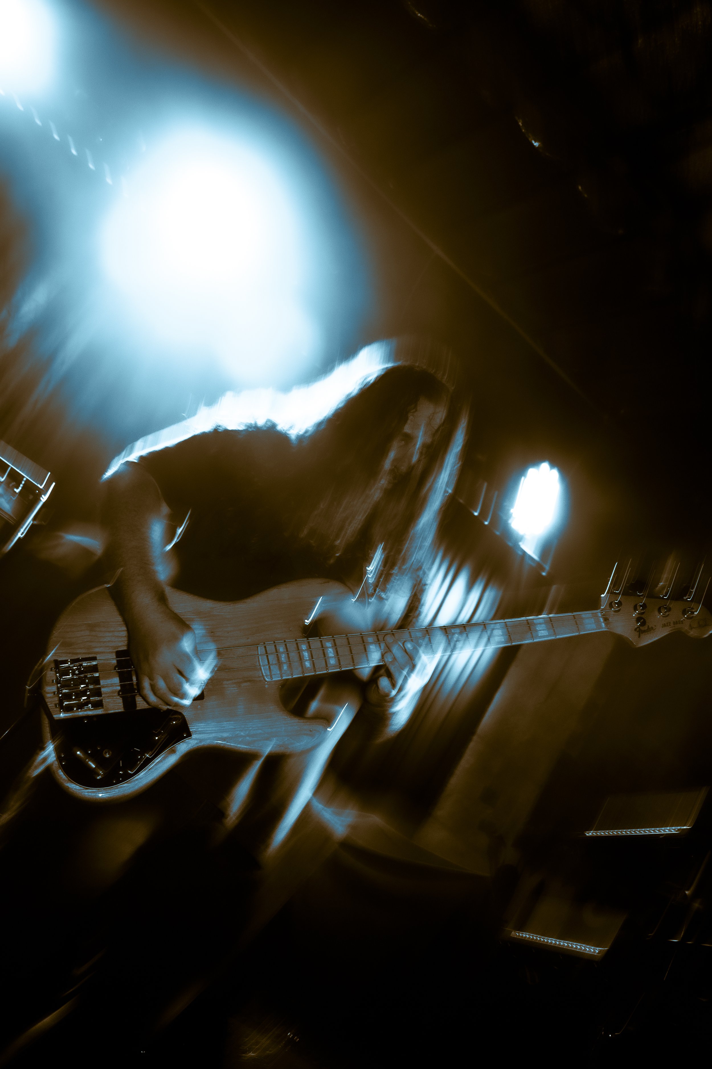 Tysonious Mink, bassist for the band LIquid Pennies with long hair playing an electric bass guitar on stage, illuminated by bright blue and warm lighting, with motion blur creating a dynamic effect.