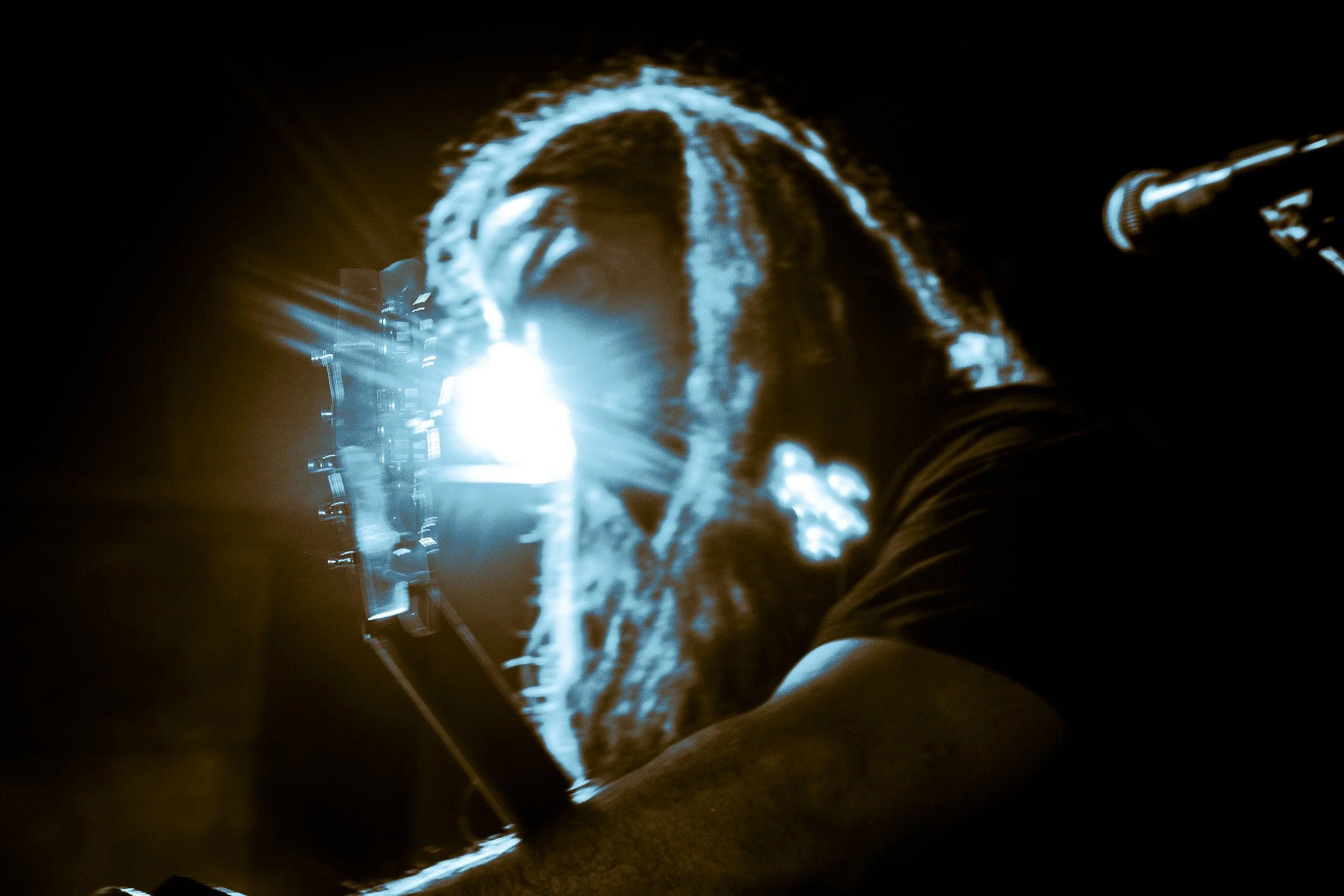 Person with brightly lit dreadlocks facing a bright light, in a dark environment, with a camera on a tripod nearby.