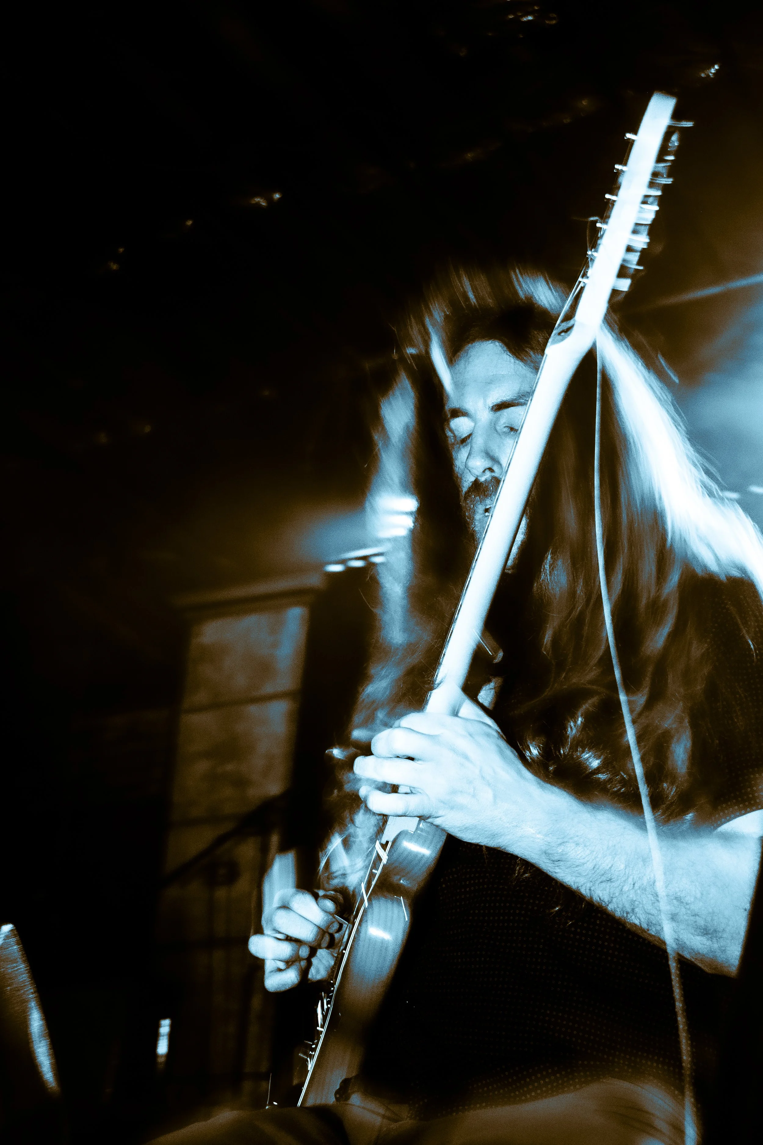 A man with long hair and a beard playing an electric guitar, photographed in blue tones with a blurred background.