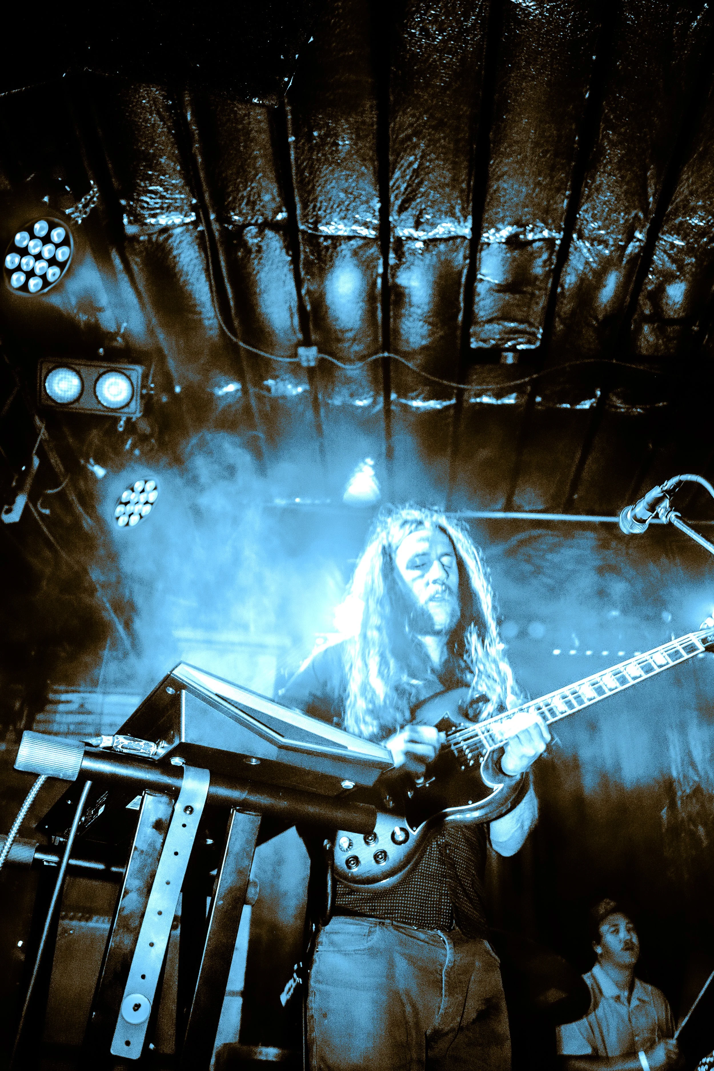 A musician with long hair playing an electric guitar on stage illuminated by blue lights, with stage equipment and a man in the background.