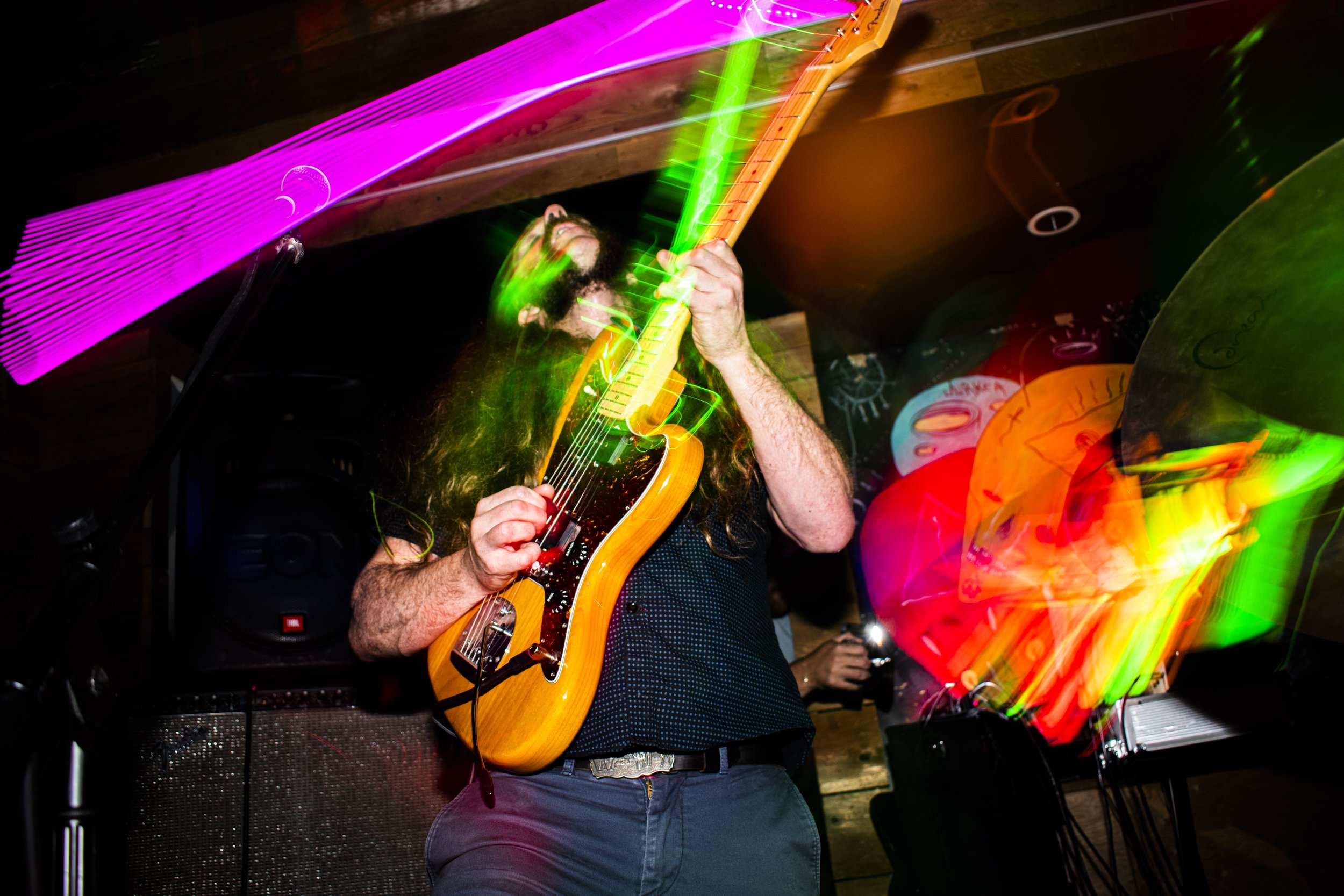Liquid Pennies frontman/guitarist Chas Binns playing an electric guitar with colorful light streaks around him in a dark room, creating a vibrant, energetic scene.
