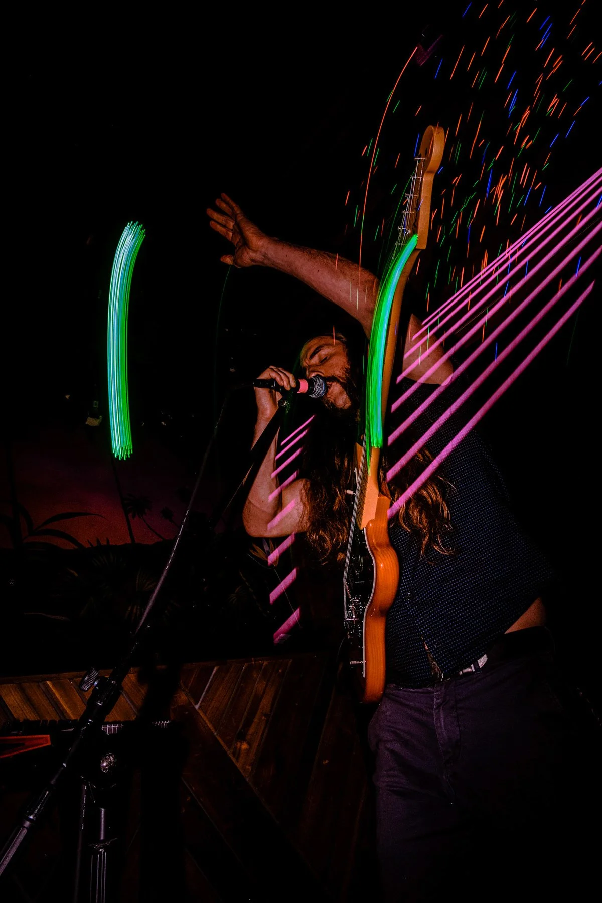 Musician with long hair and beard singing into a microphone with a guitar on his chest, surrounded by colorful light streaks during a live performance at night.