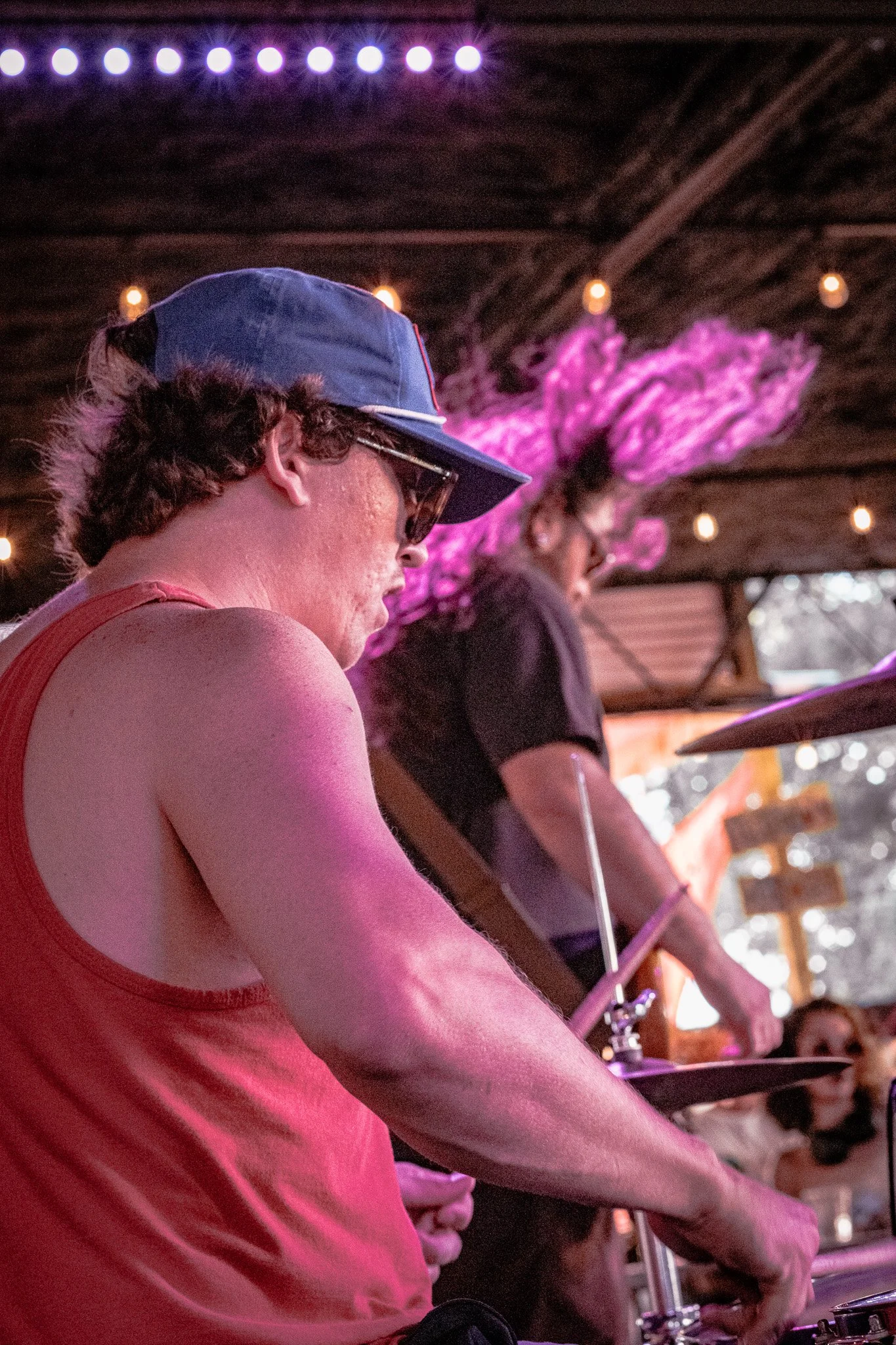 Liquid Pennies drummer Pierson Whicker with a blue cap, sunglasses, and a red tank top performing at a music event under purple lights, with a woman with vibrant pink hair in the background.