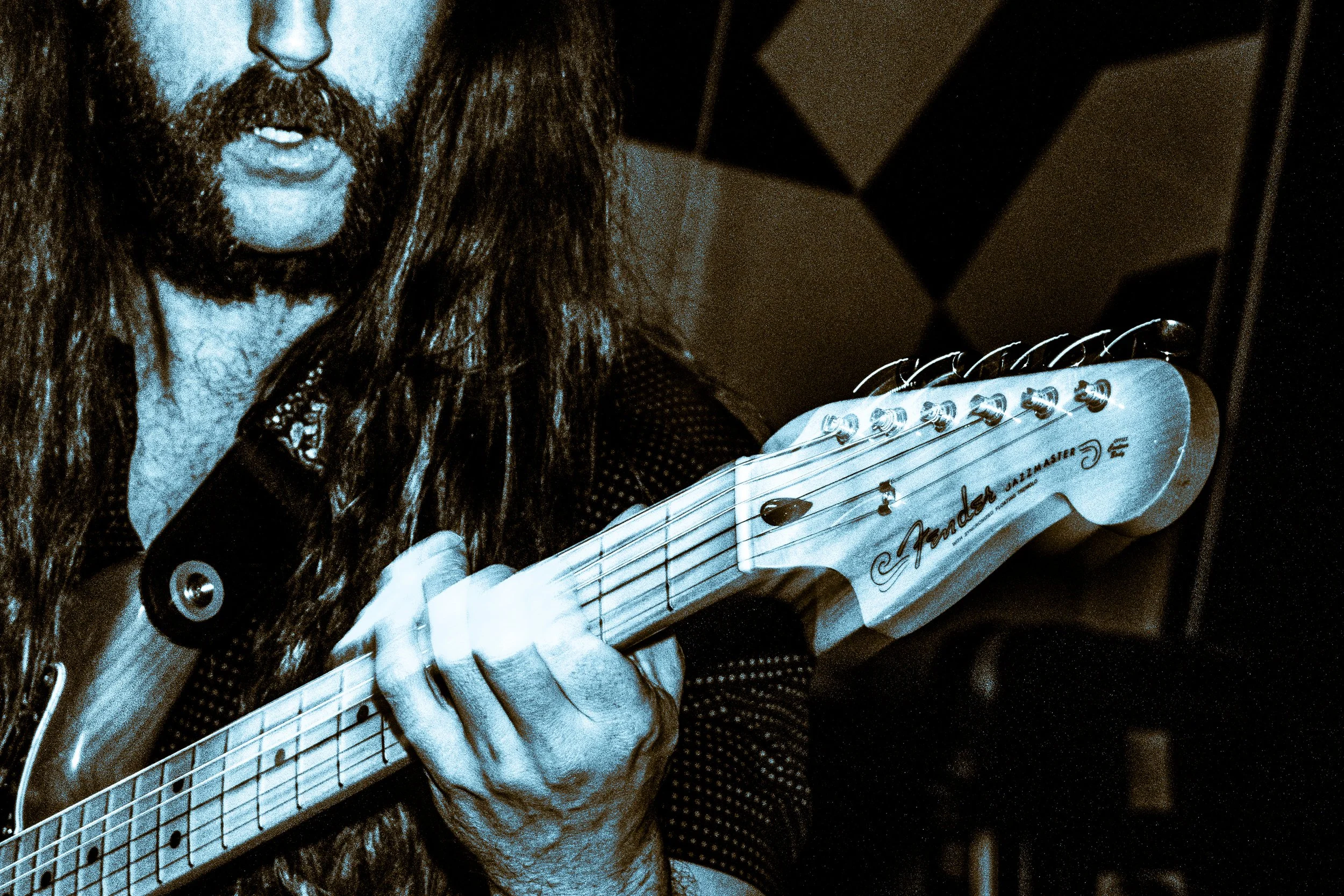 Close-up of a person with long hair and beard playing an electric guitar, with focus on the guitar's headstock and hand.