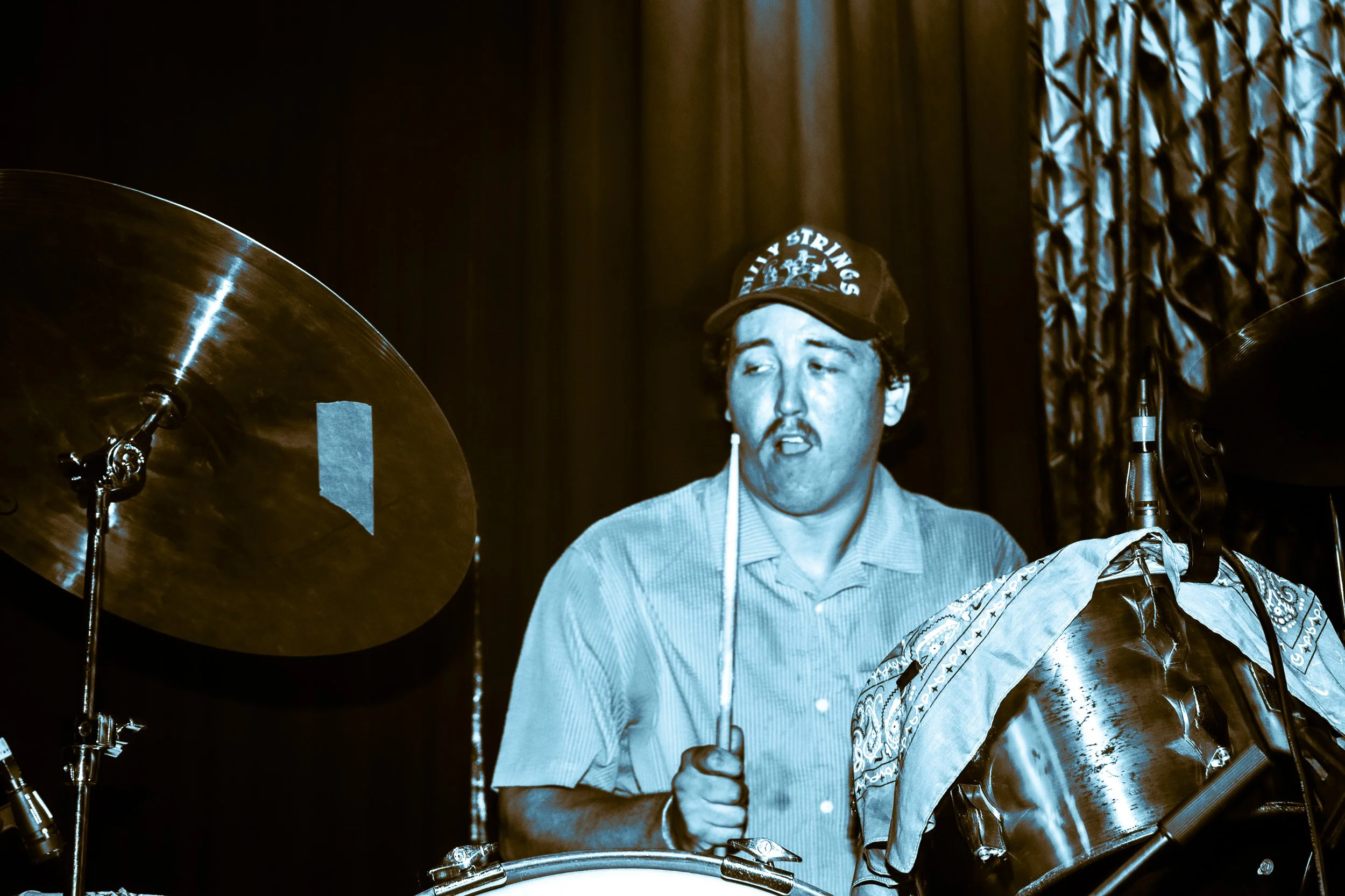 A man with a mustache wearing a cap and a striped shirt playing drums on stage, with a curtain in the background.