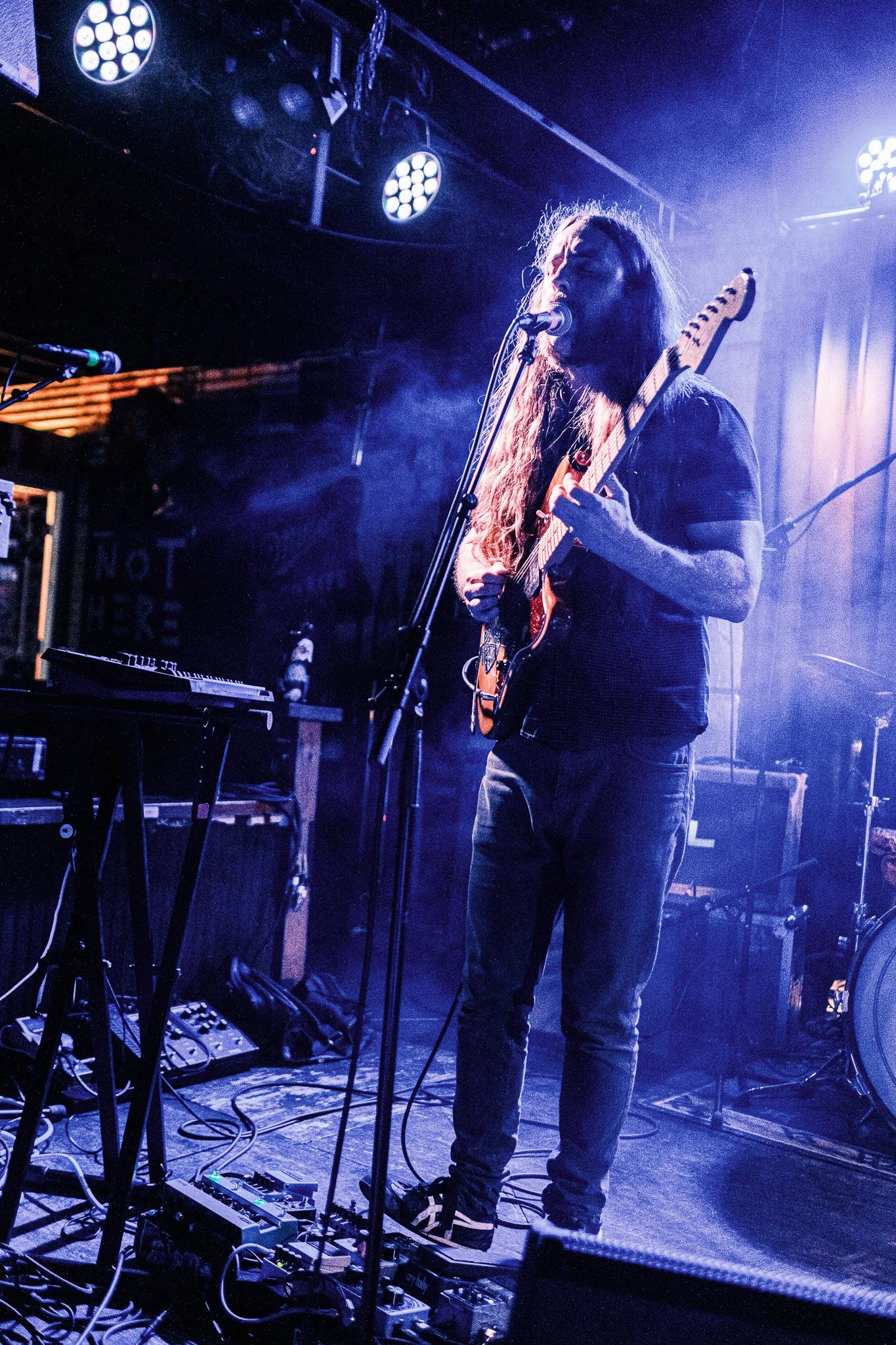 A musician with long hair is performing on stage, playing an electric guitar and singing into a microphone, surrounded by stage lights and musical equipment.