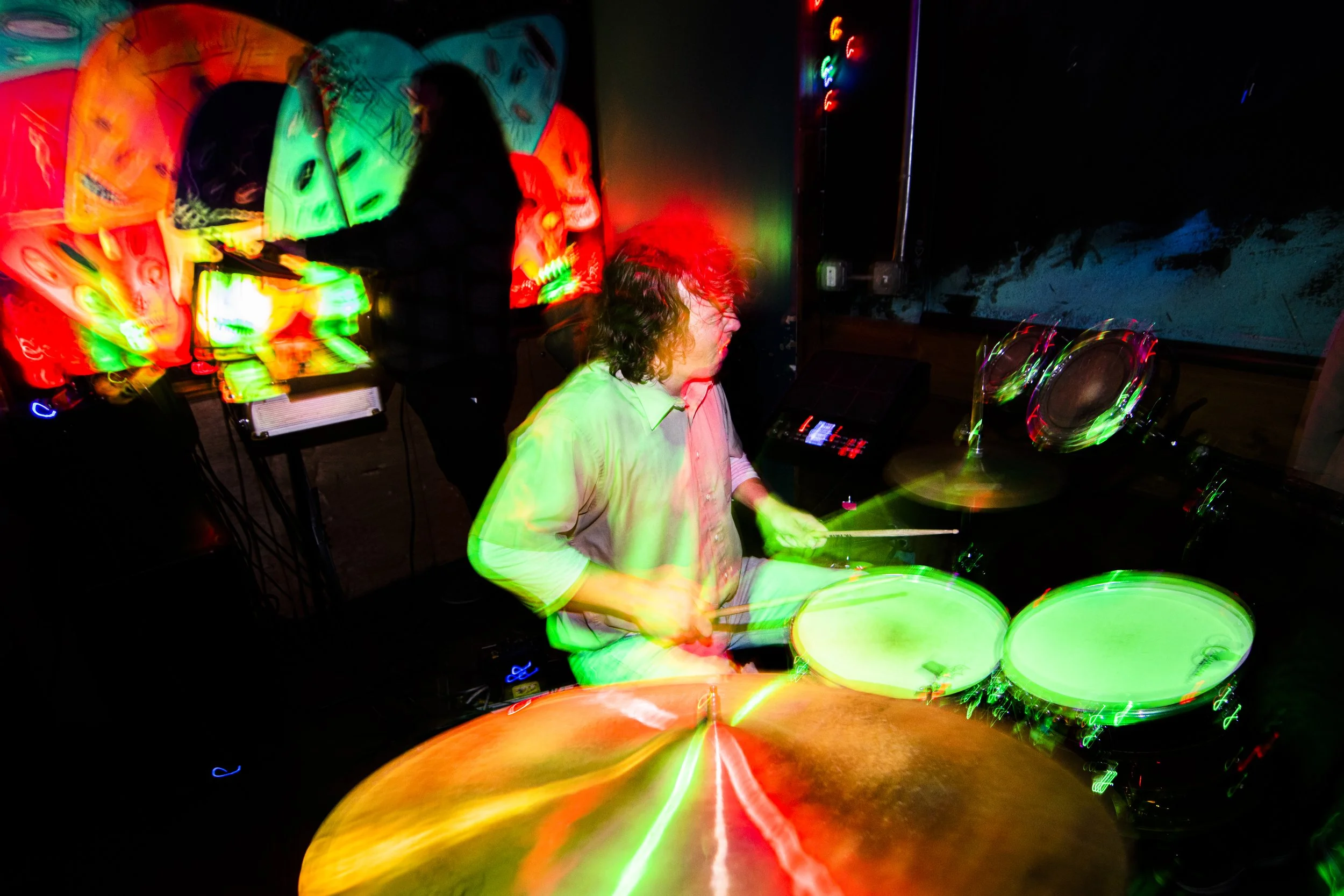 Person playing drum set in a dimly lit room with colorful, blurred light effects.
