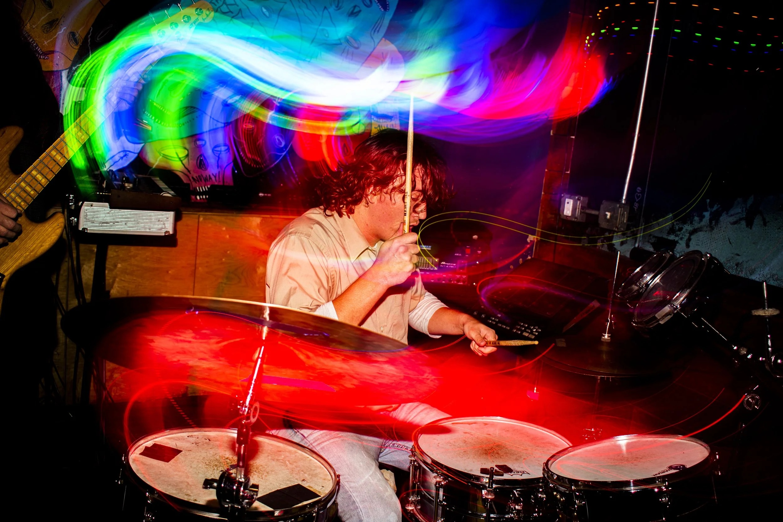 Liquid Pennies drummer Pierson Whicker playing drums and Tysonious Mink playing bass in a dimly lit room with colorful light streaks due to long exposure photography.
