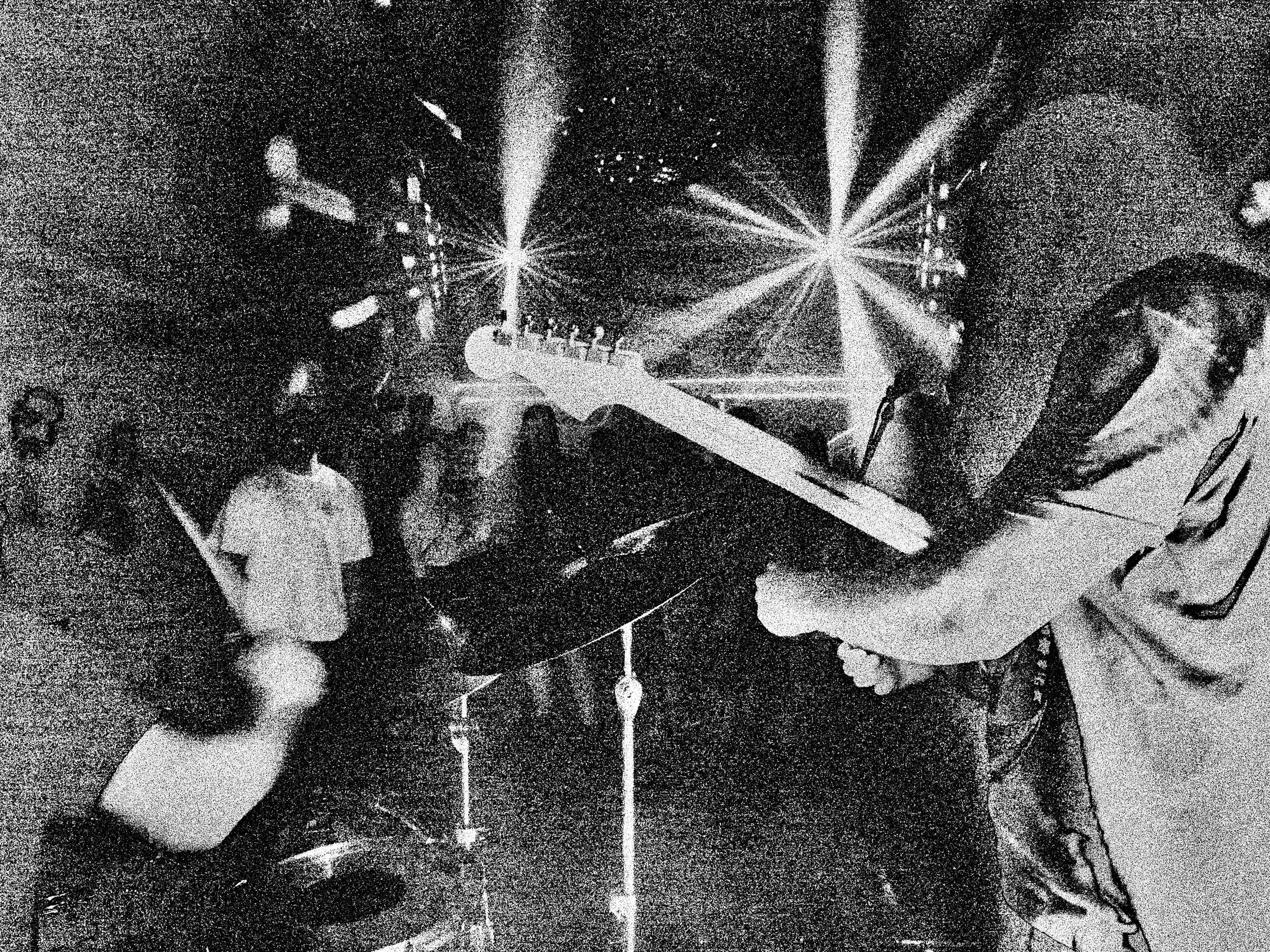A DJ wearing glasses and headphones operates a turntable, while a woman with curly hair and a white top stands nearby, both under bright stage lights in a dark club setting.