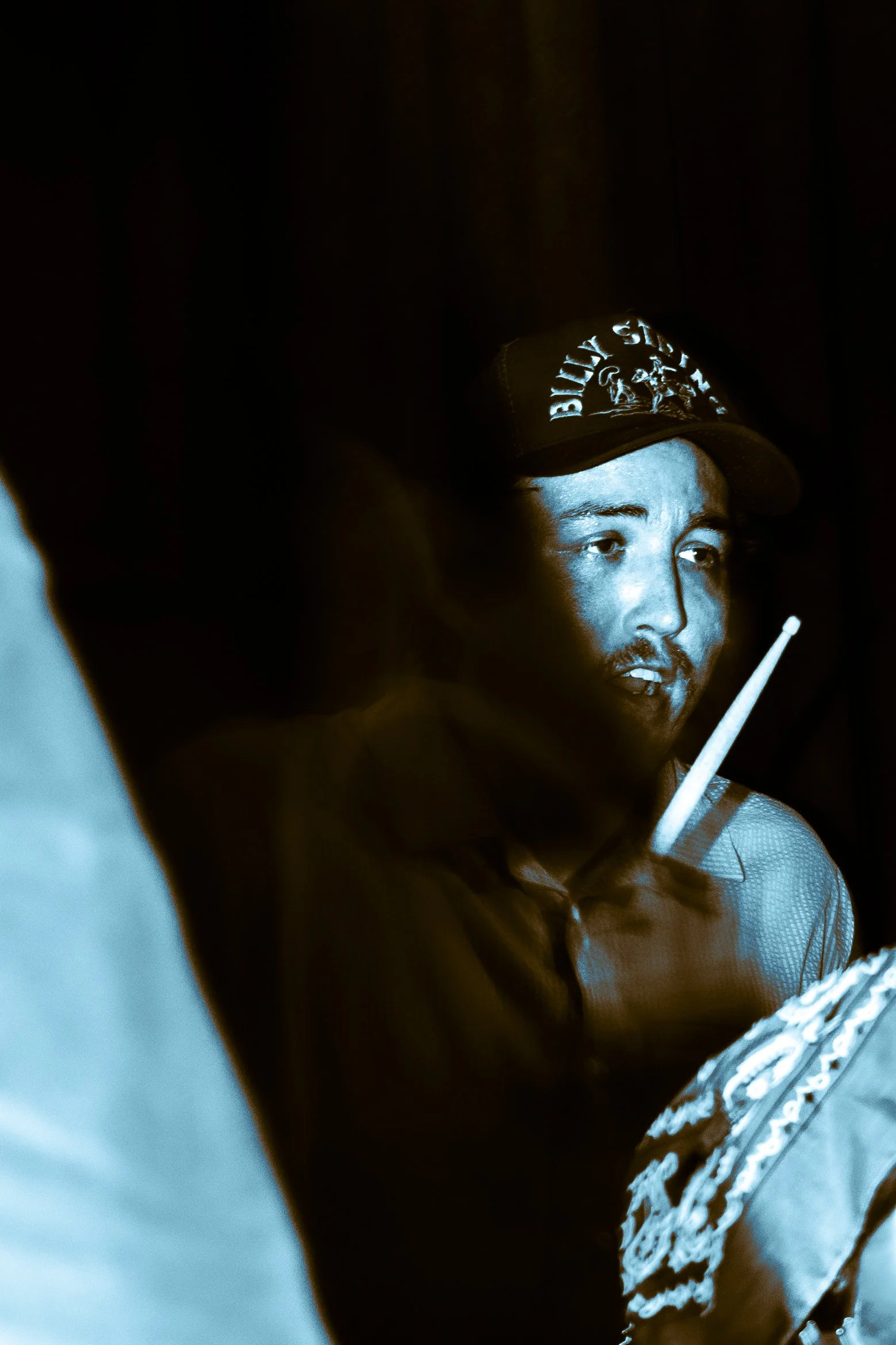 A man wearing a cap with the words 'BEYER SIGN' illuminated by a blue light, holding a cigarette, with a dark background.