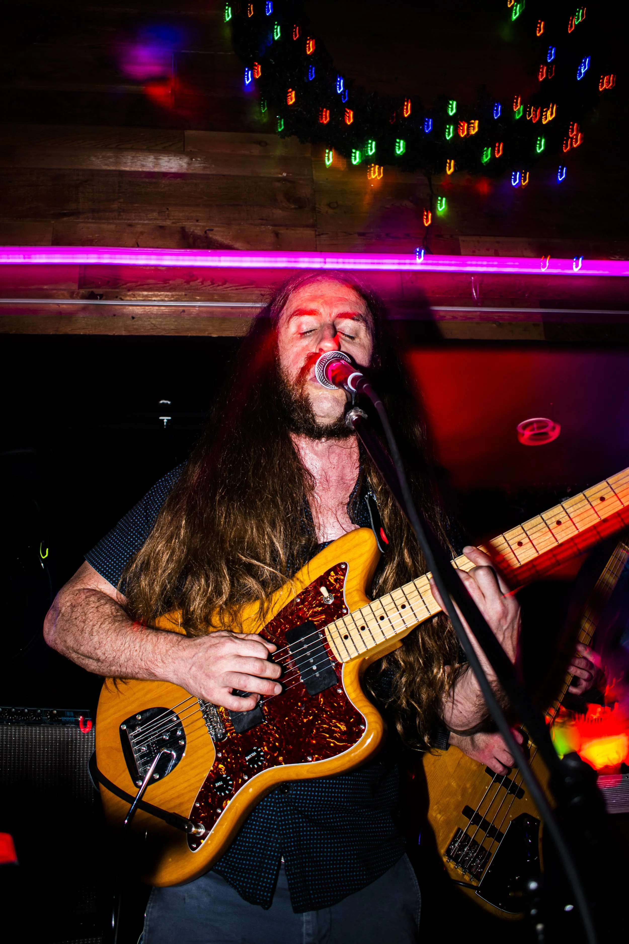 Liquid Pennies frontman/guitarist Chas Binns playing an electric guitar and singing into a microphone in a dimly lit venue with colorful, blurred lights in the background.
