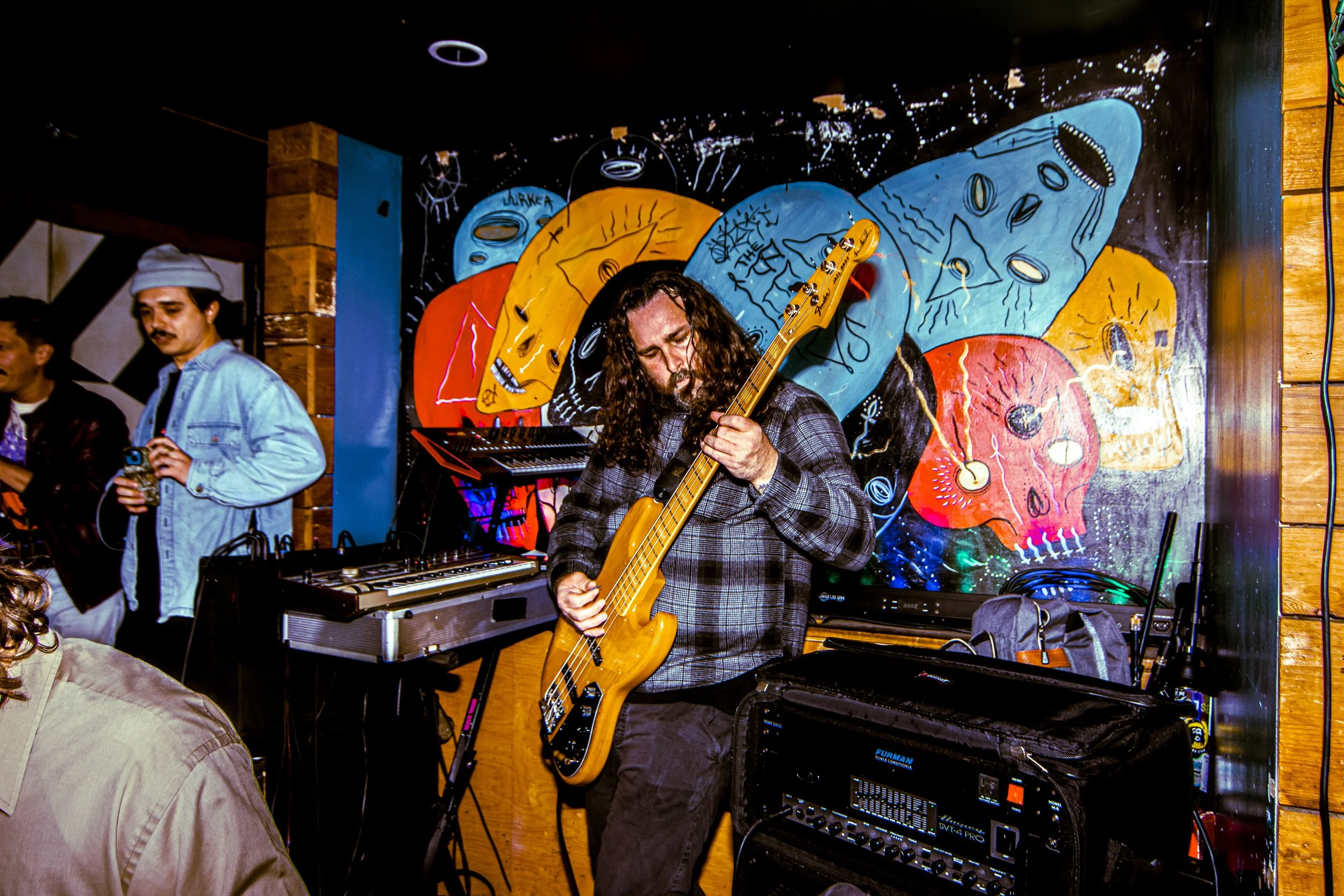 Musicians performing in front of a colorful skull mural, with one playing an electric bass guitar and another using electronic music equipment.