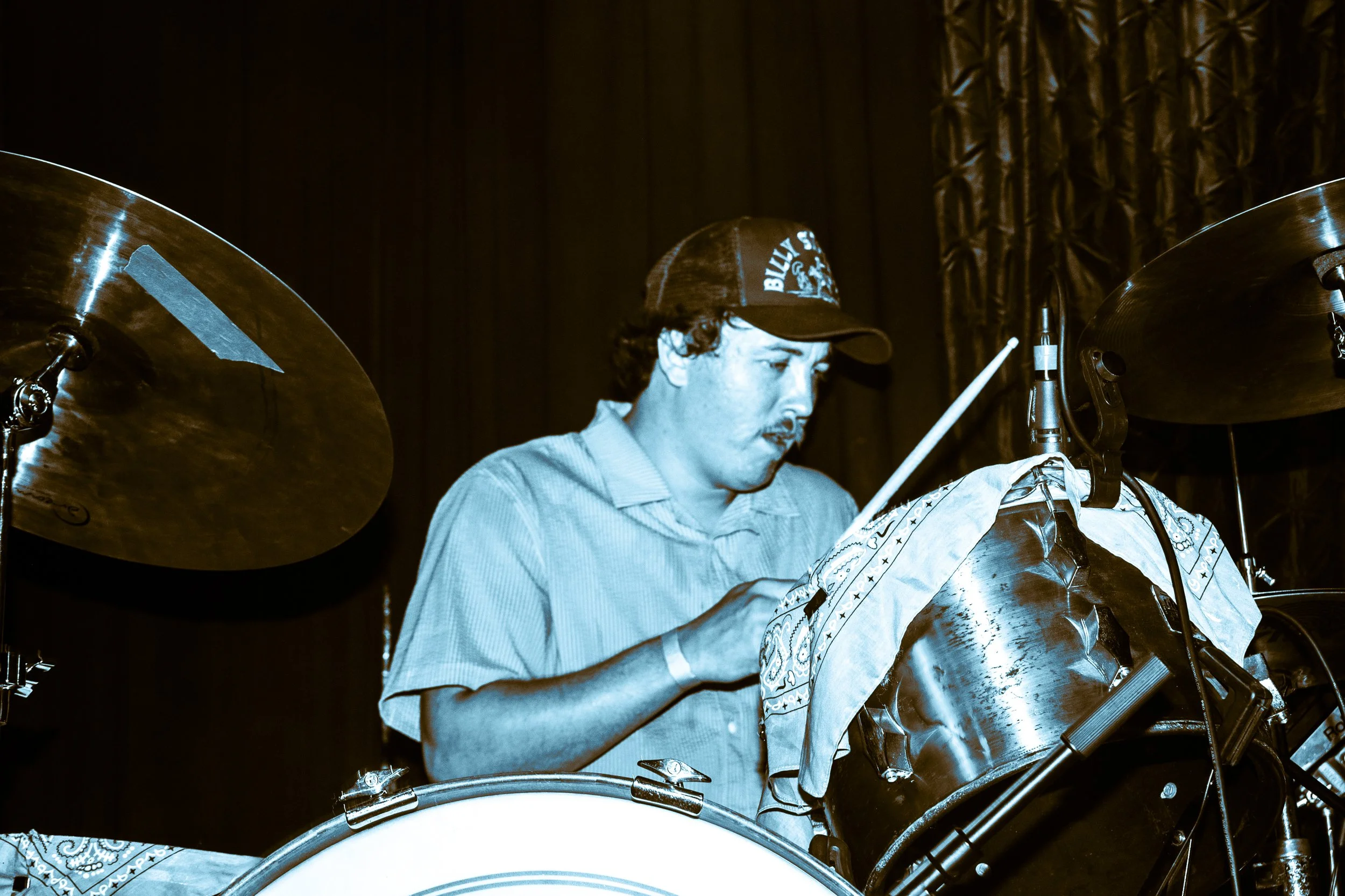 Psych rock band Liquid Pennies drummer PIerson Whicker wearing a cap and focused on his drumming, with dark curtains in the background.