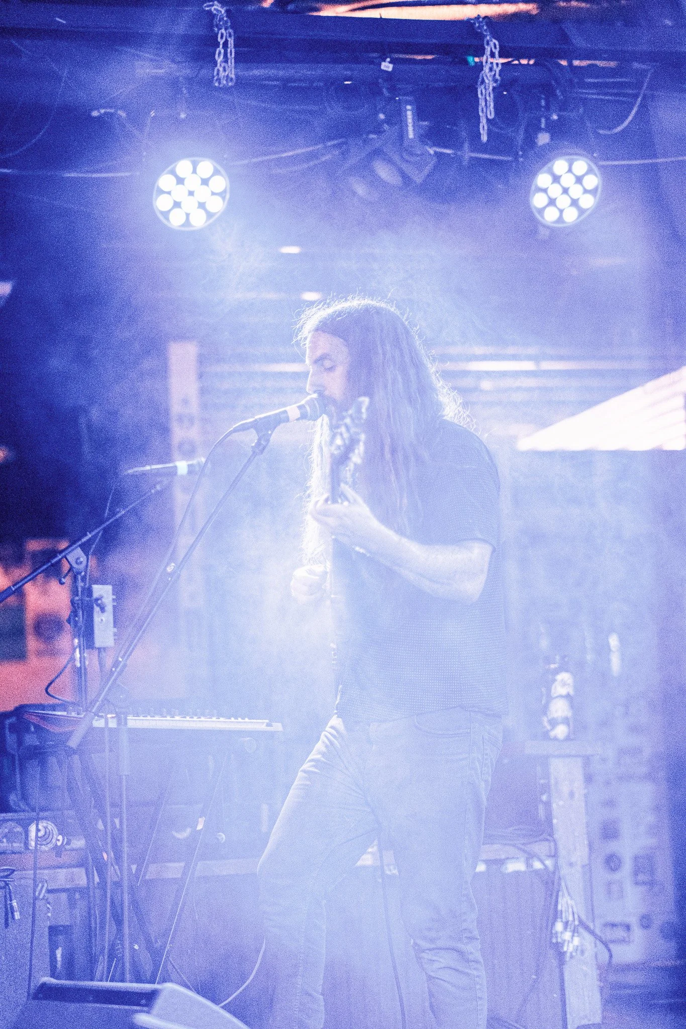 A person with long hair singing into a microphone while playing a guitar on stage, illuminated by blue stage lights.