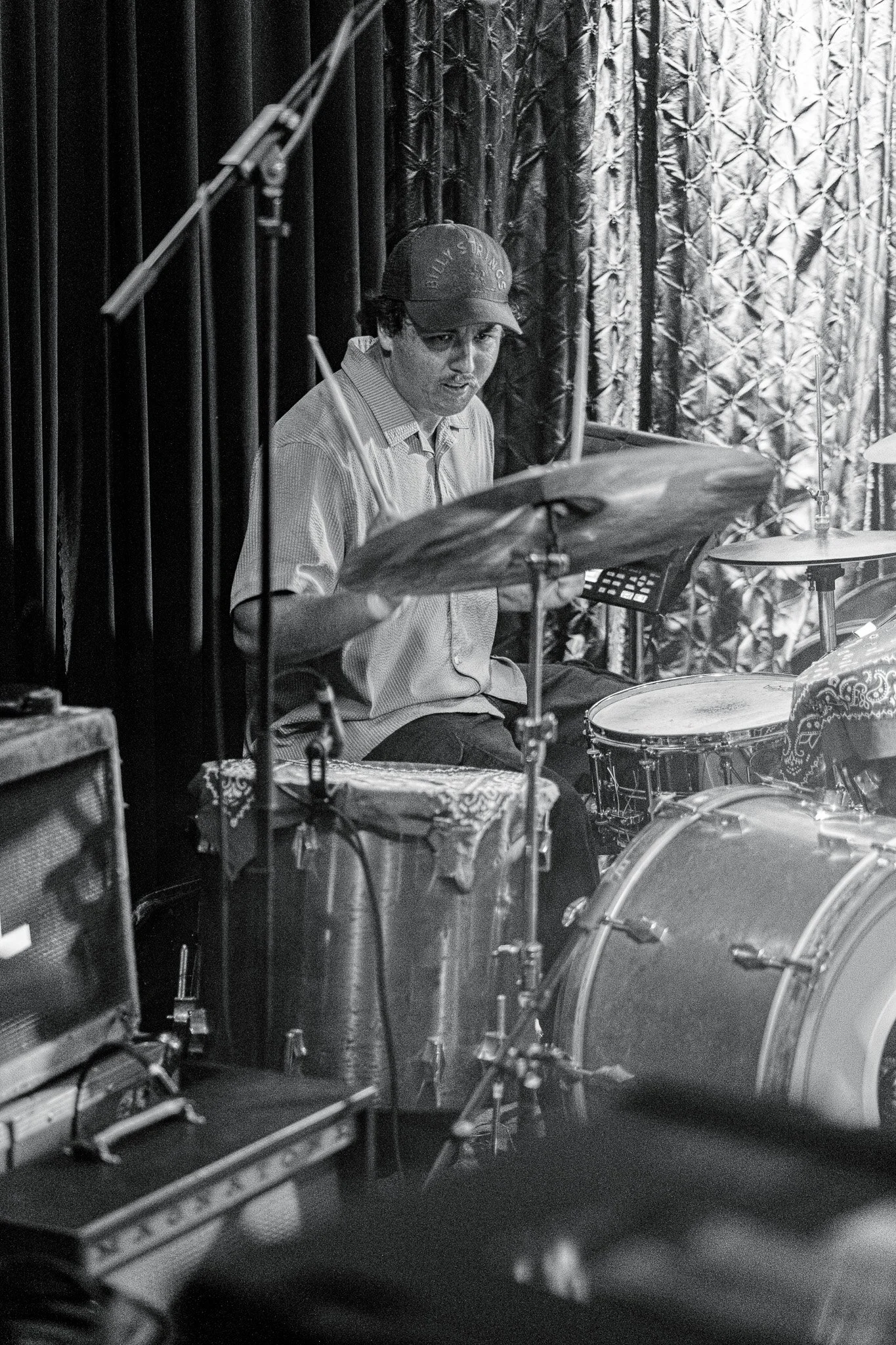 A man playing drums in a recording studio or live performance setting, with soundproofing curtains in the background.