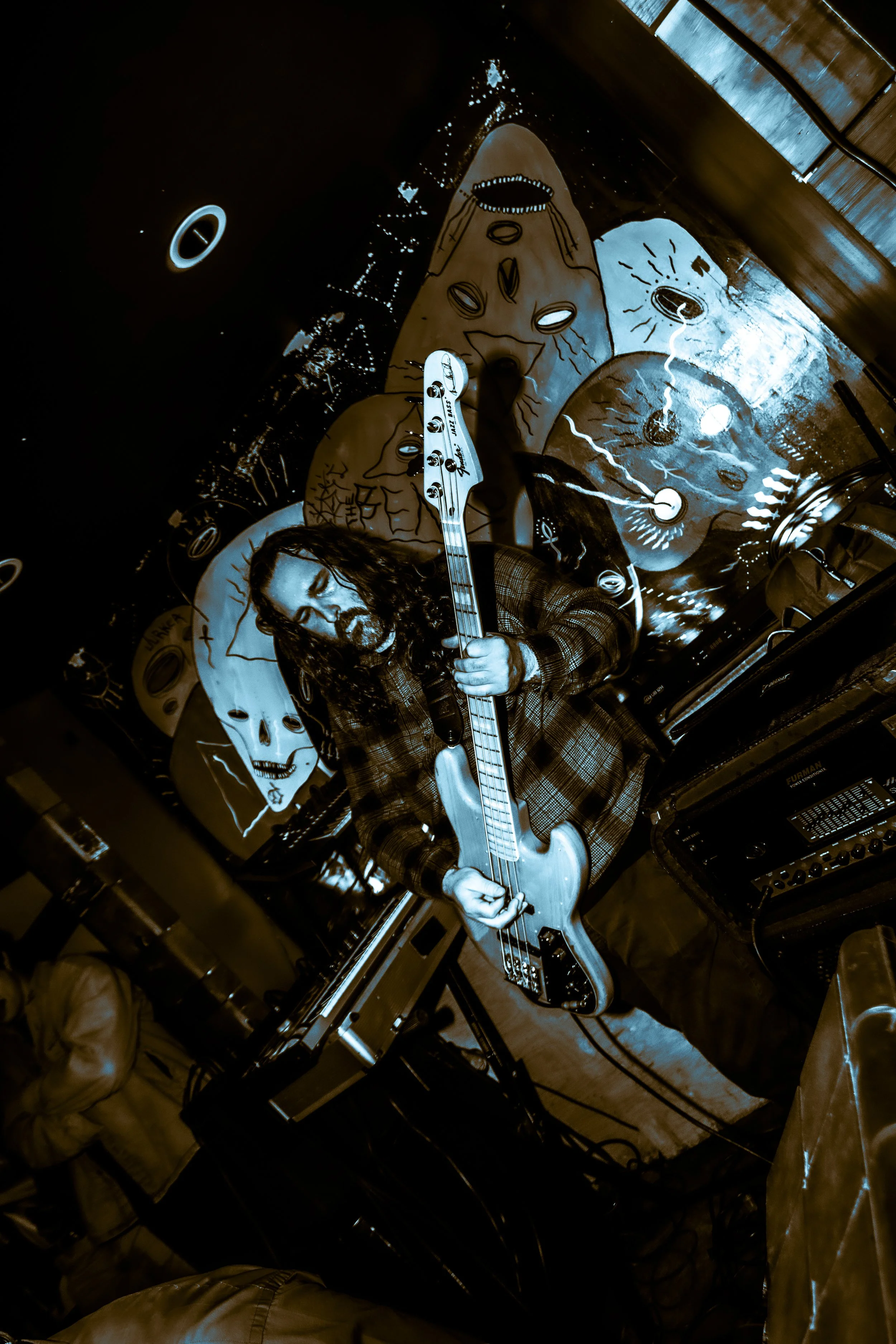 A man with long curly hair and a beard playing an electric bass guitar in a dimly lit room with abstract art and skull images on the wall behind him.