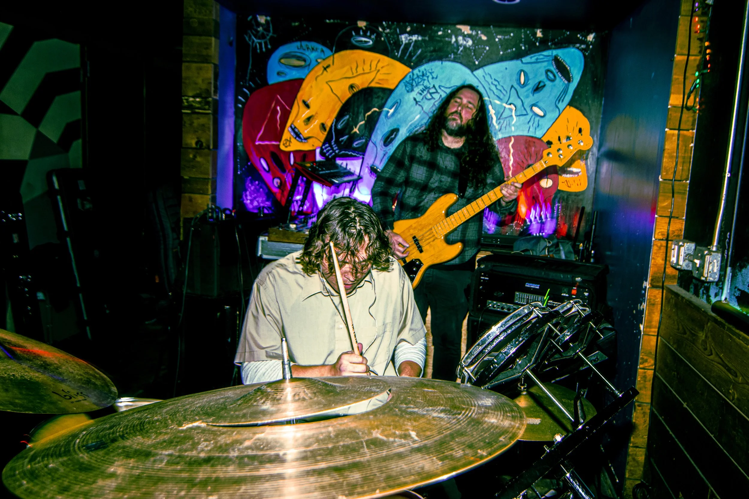 Two musicians performing in a dimly lit venue with a colorful mural of cartoon-like faces behind them; one playing drums with a drumstick in hand, and another playing bass guitar with a colorful mural of cartoon-like faces behind them.