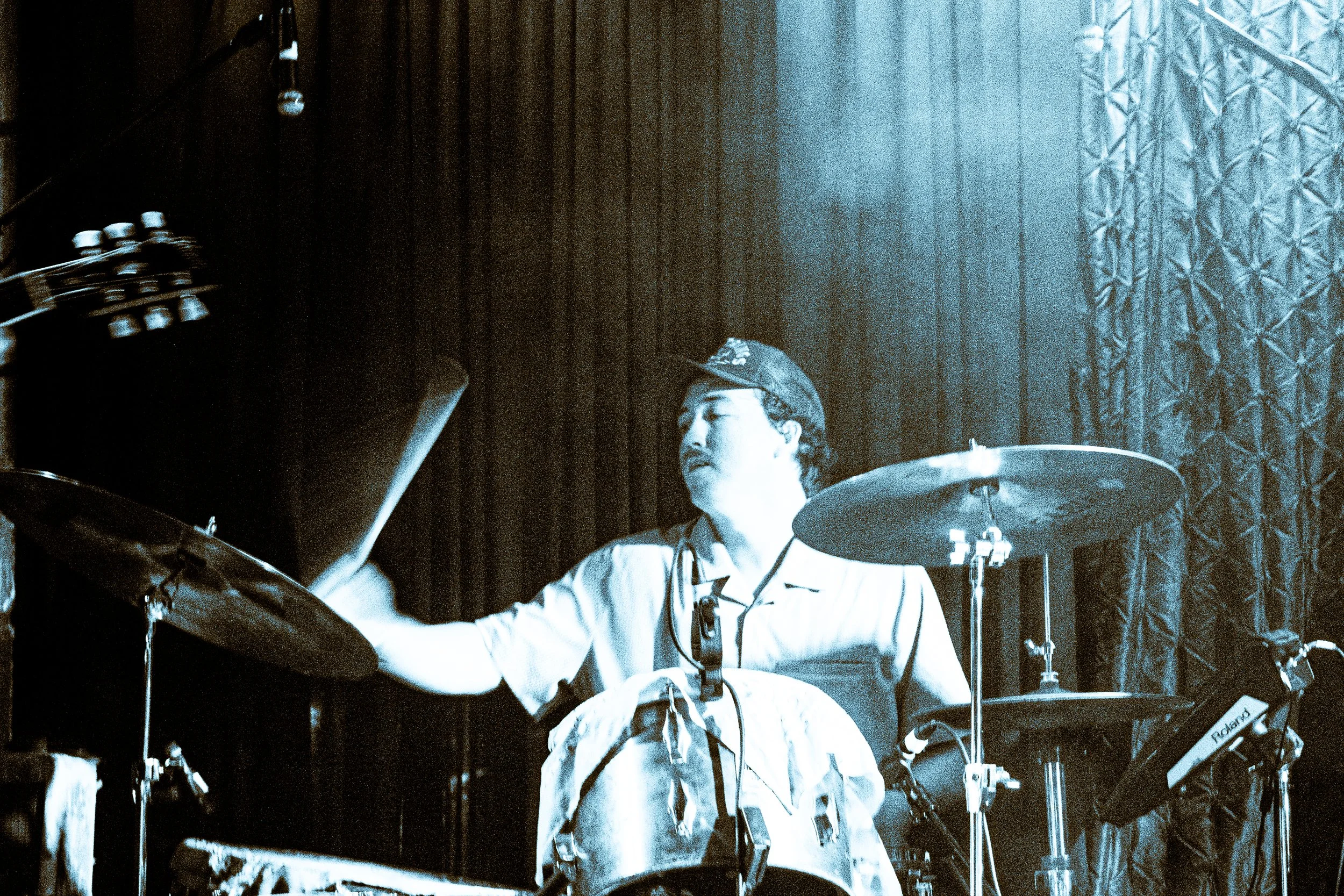 Psych rock band Liquid Pennies drummer PIerson Whicker playing a drum set on stage, wearing a backward cap, light-colored shirt, and headphones, with a dark curtain background.
