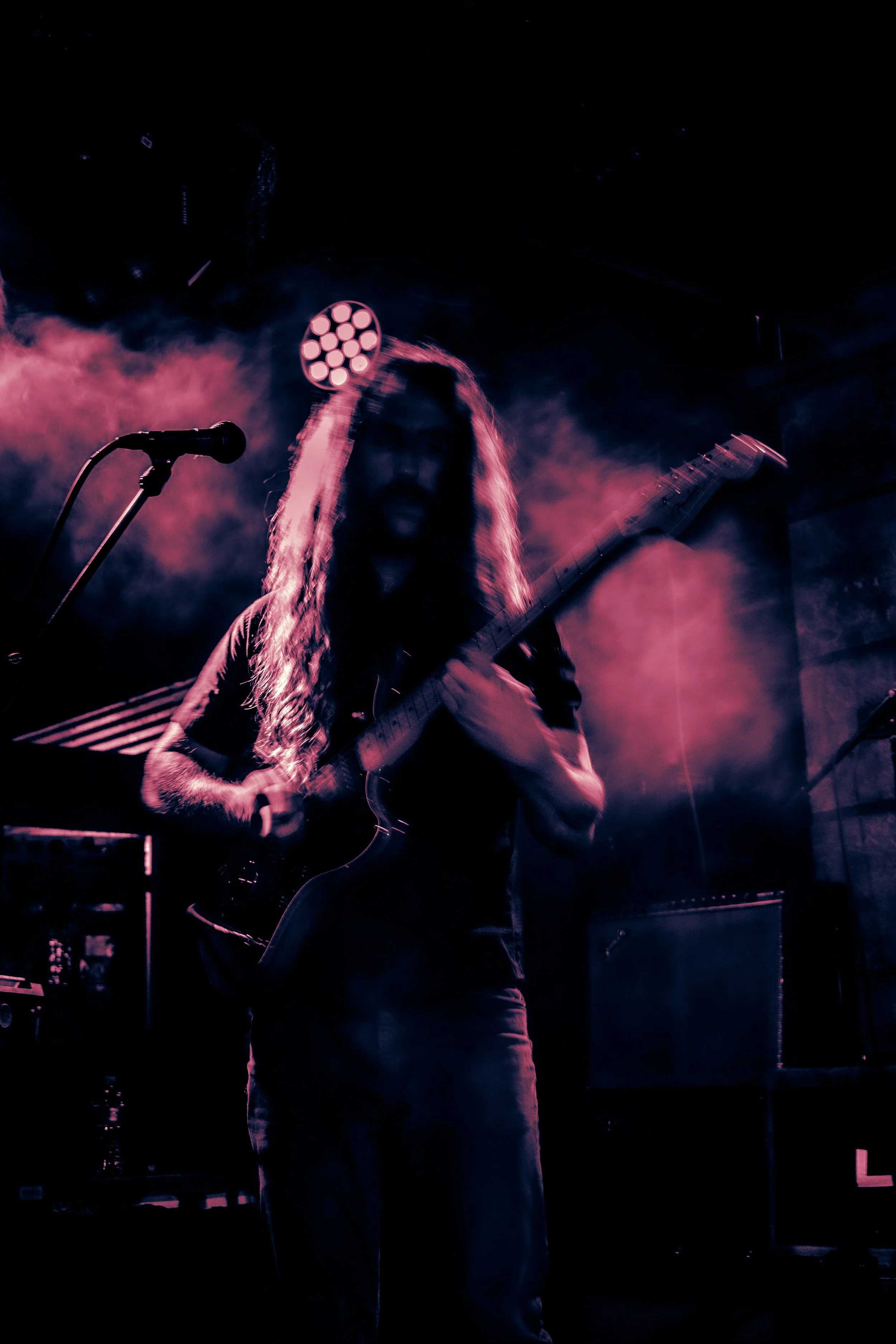 Rock band Liquid Pennies frontman/guitarist Chas BInns playing an electric guitar on stage with purple and pink lighting and smoke in the background.