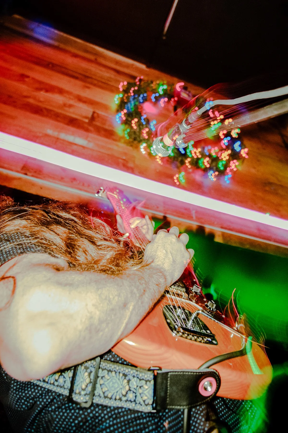 Person playing electric guitar in a room decorated with Christmas lights and wreaths, with neon lighting.