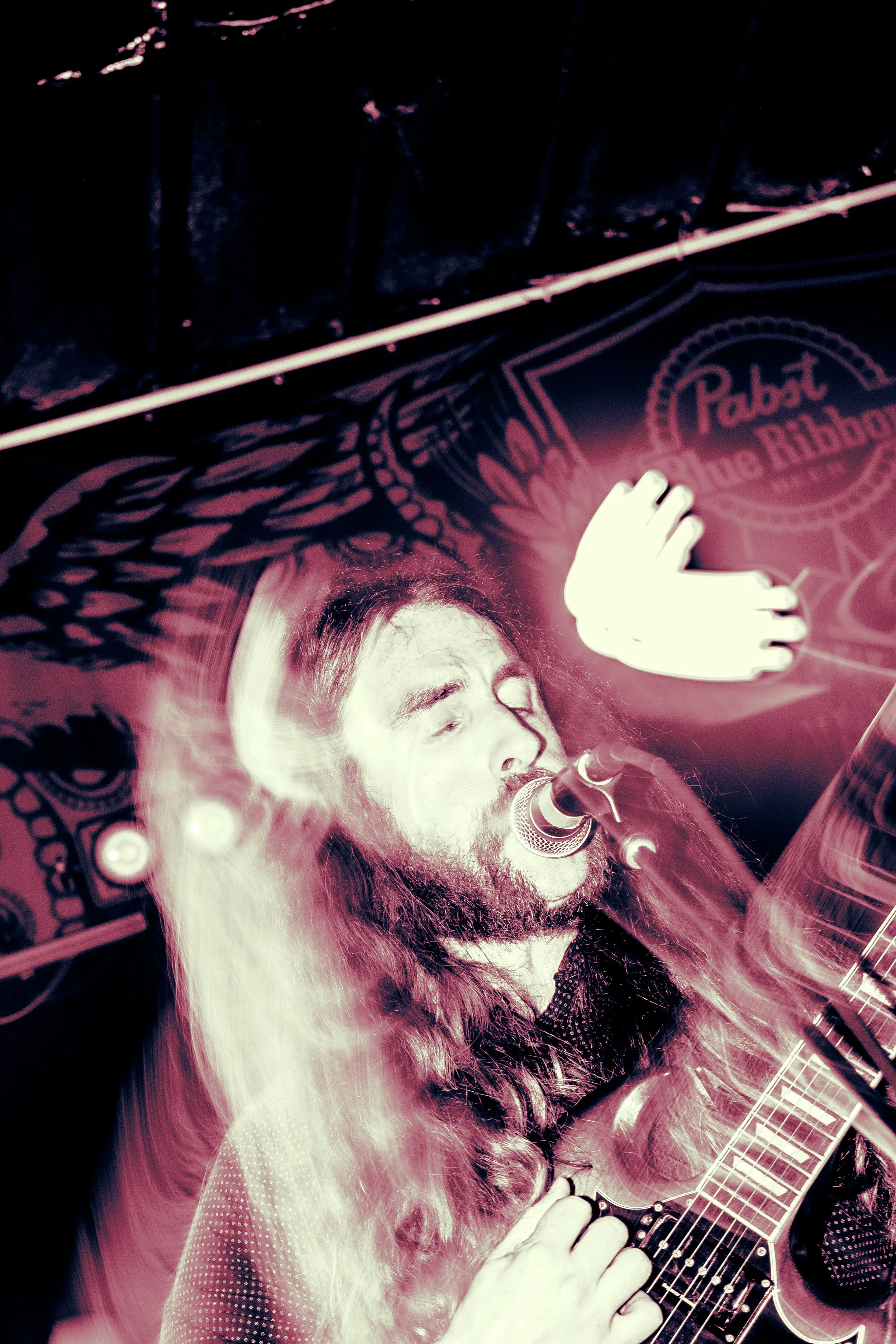 Rock band Liquid Pennies frontman/guitarist Chas BInns, a male singer with long hair and a beard passionately singing into a microphone while playing a Gibson SG electric guitar, in front of a background featuring a "Pabst Blue Ribbon" banner.