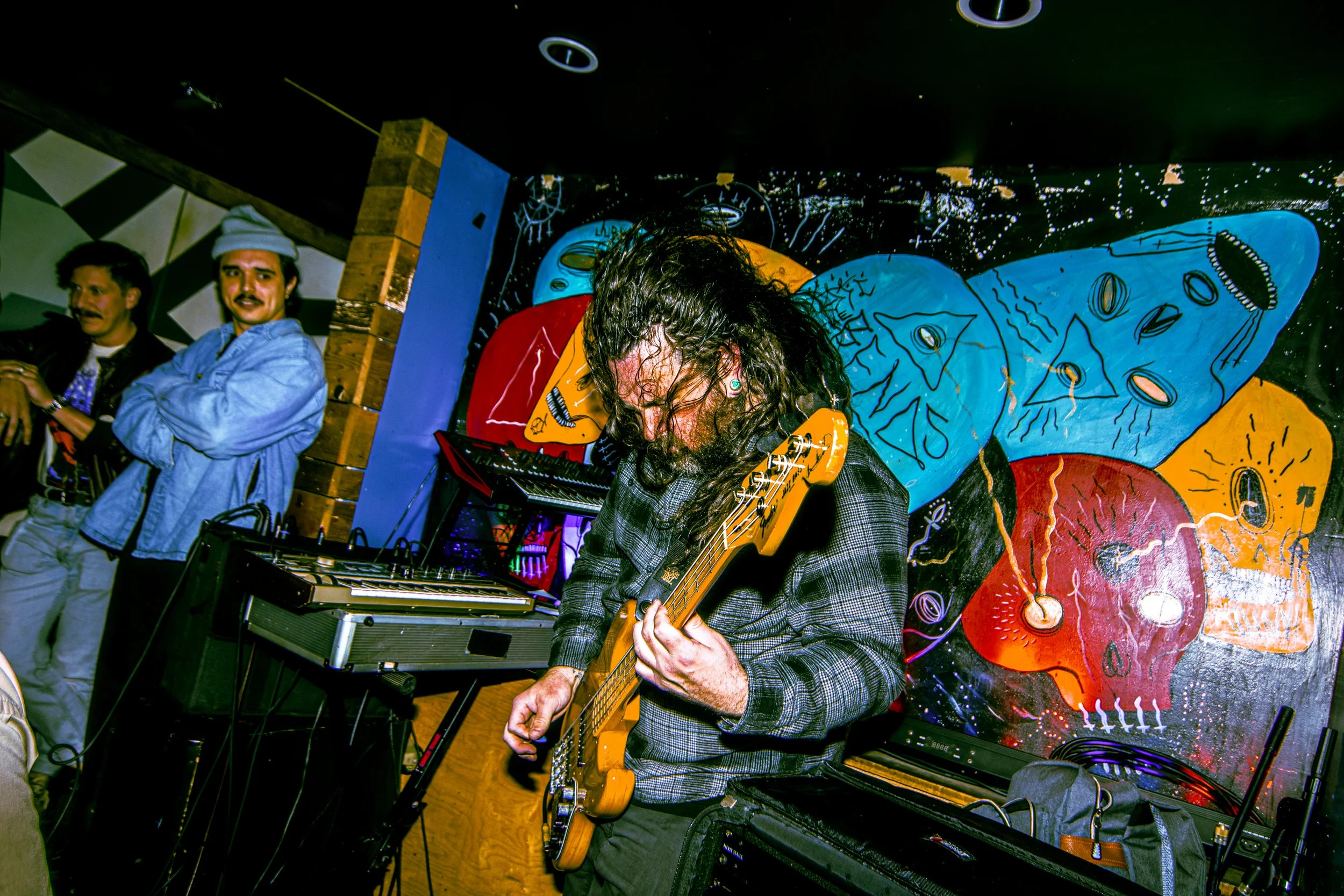 Musician playing electric guitar in front of colorful, abstract mural during a live music performance at a lively venue.