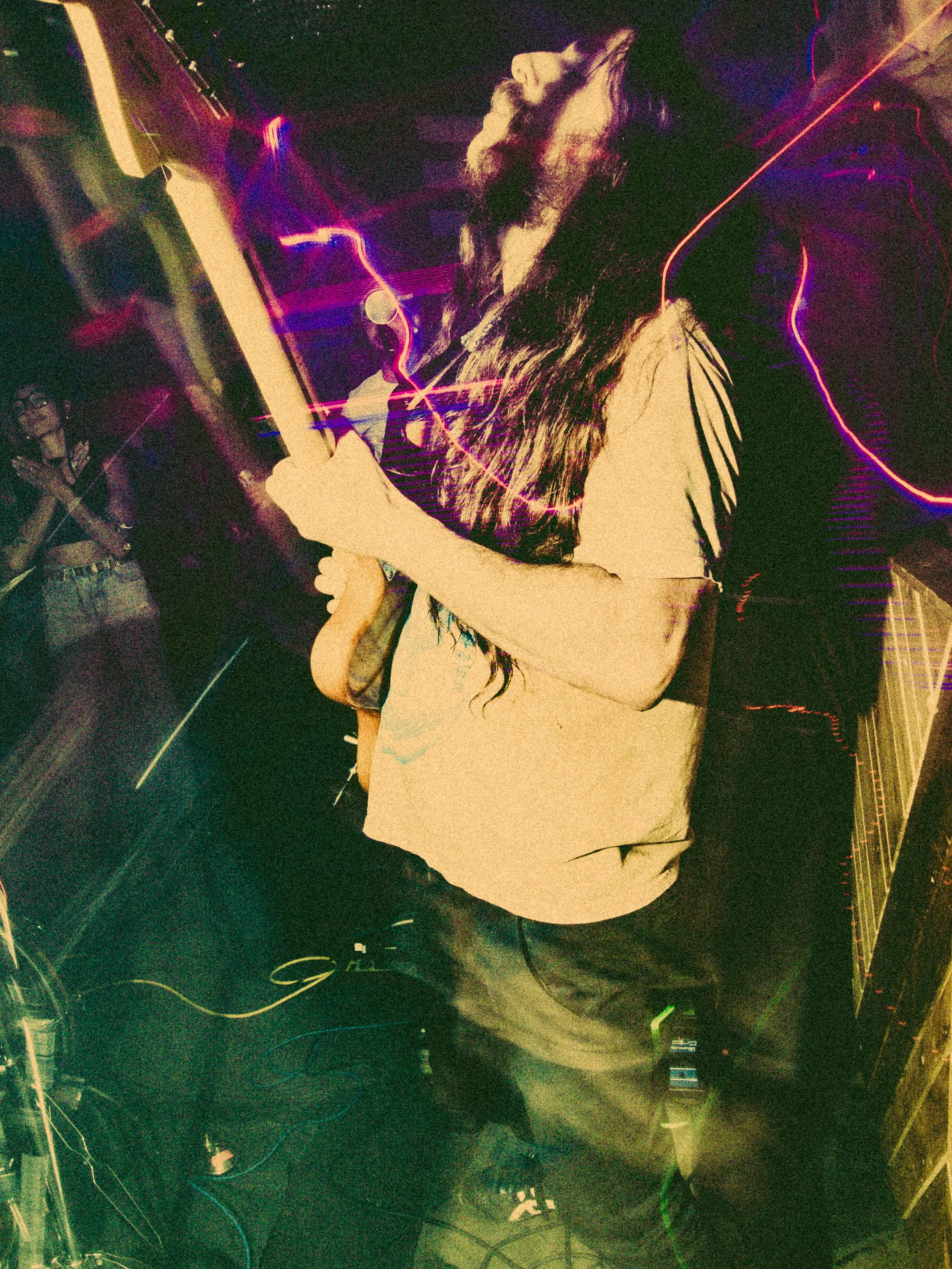 A person with long dark hair and a light-colored shirt is DJing at a nightclub, with colorful light streaks in the background.