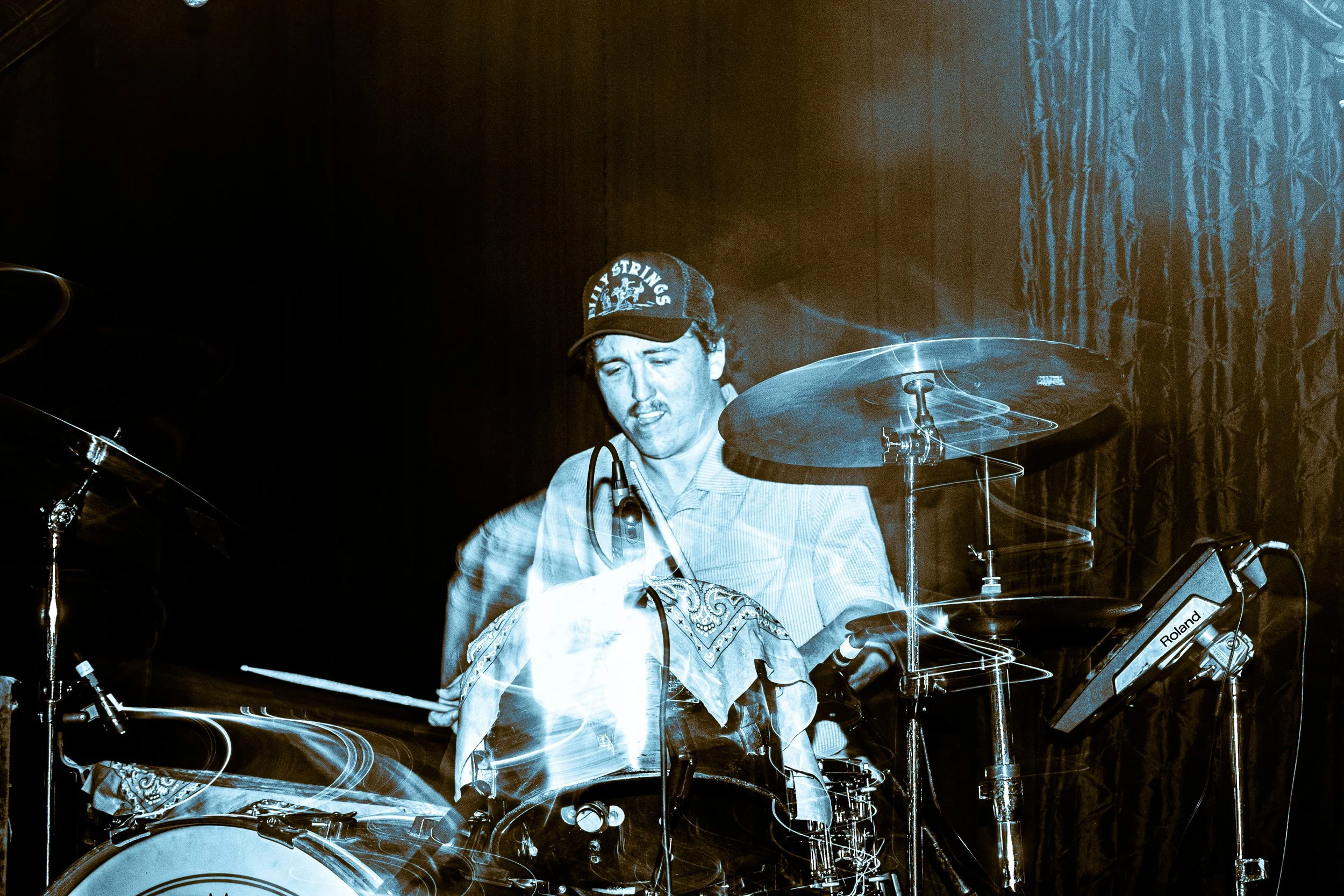 Psych rock band Liquid Pennies drummer PIerson Whicker  playing drums on stage, wearing a cap, with musical equipment and a dark curtain in the background.