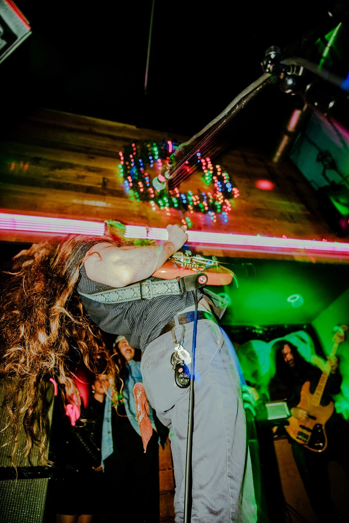 Musician with long curly hair playing guitar on stage, surrounded by colorful lights, crowd in the background