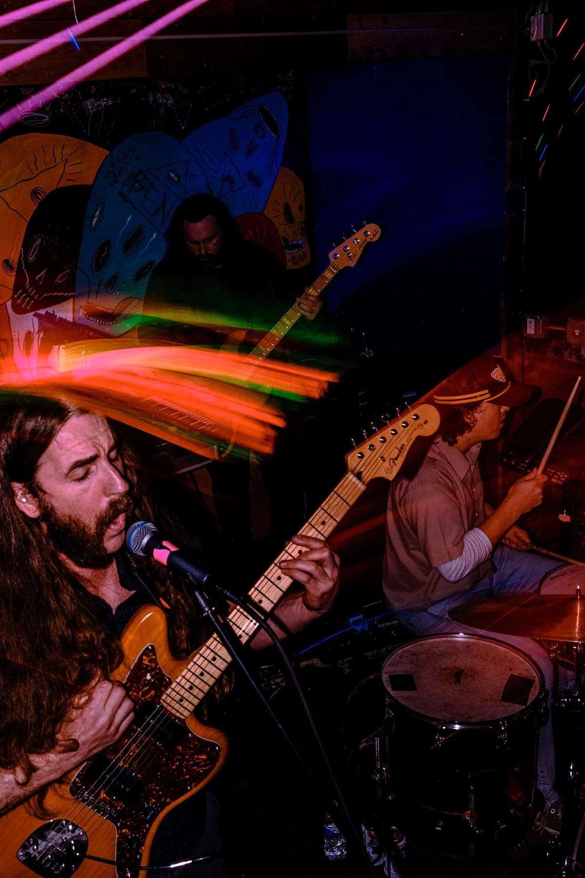 A band performing live with a guitarist singing into a microphone, a drummer playing with a stick, a bassist playing, and colorful light trails in a dimly lit venue with abstract artwork background.