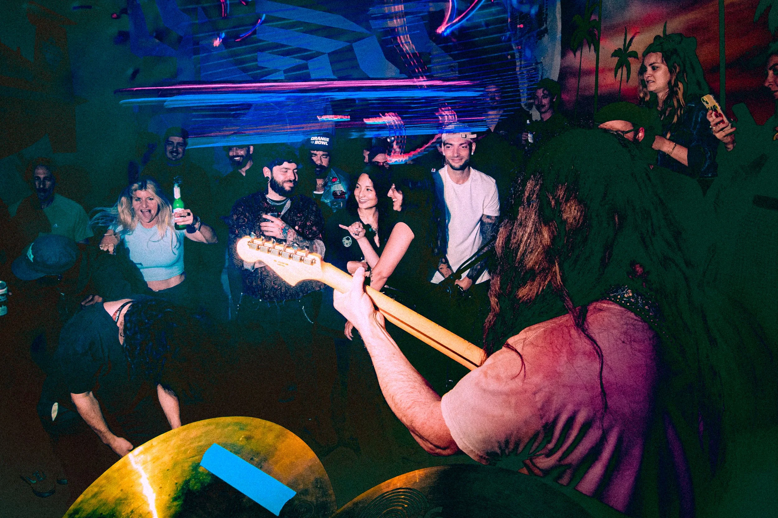 A musician with long, dark hair and a beard playing an electric guitar in front of a crowd at a lively indoor party or concert, with colorful lighting and smiling people enjoying the performance.