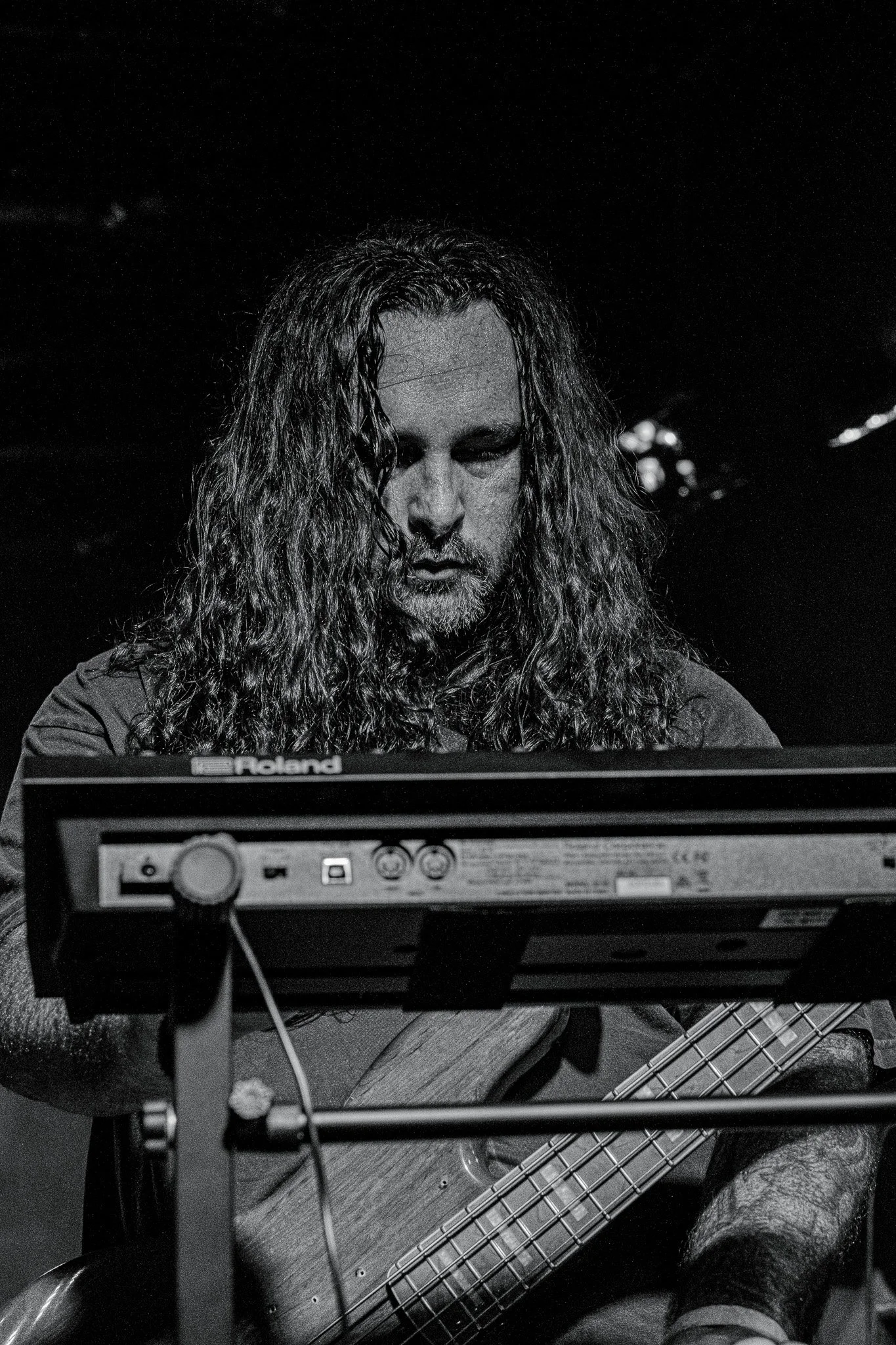 A man with long curly hair playing a bass guitar, looking down, with electronic music equipment in front of him in a dark setting.