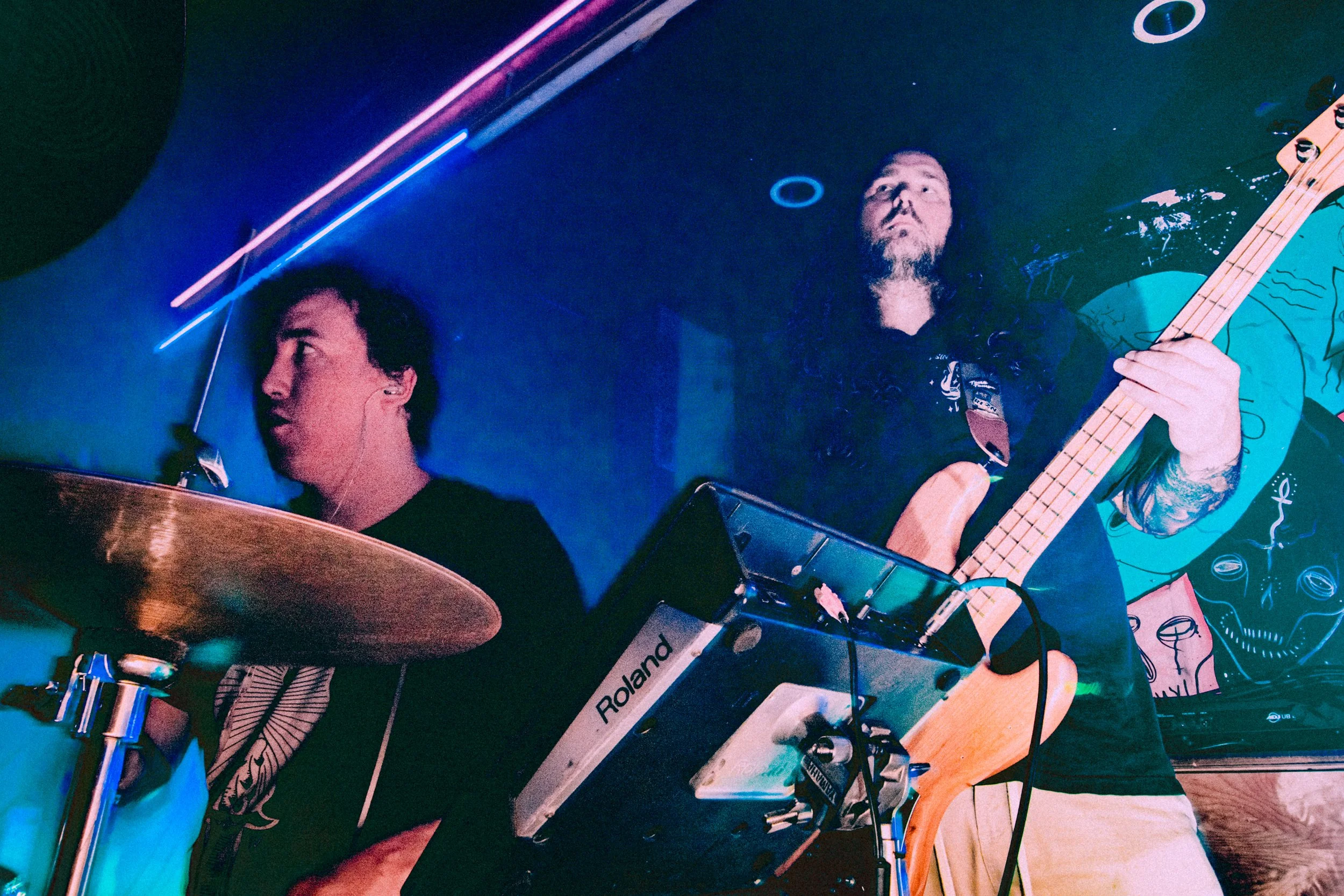 Two musicians performing live; a man on the left is playing drums and a man on the right is playing a bass guitar.