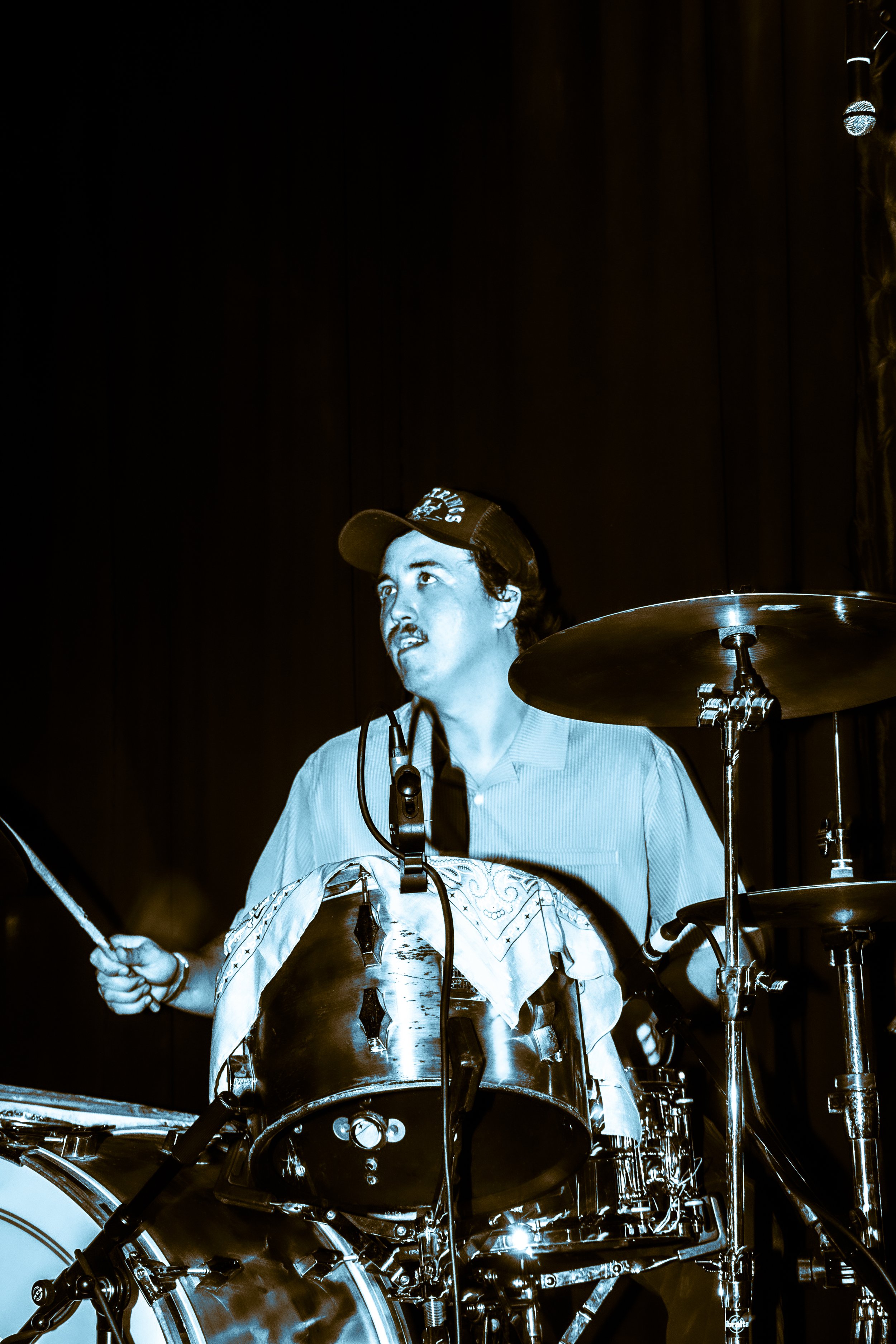 Musician playing drums on stage, wearing a baseball cap and shirt, captured in black-and-white with a blue hue.
