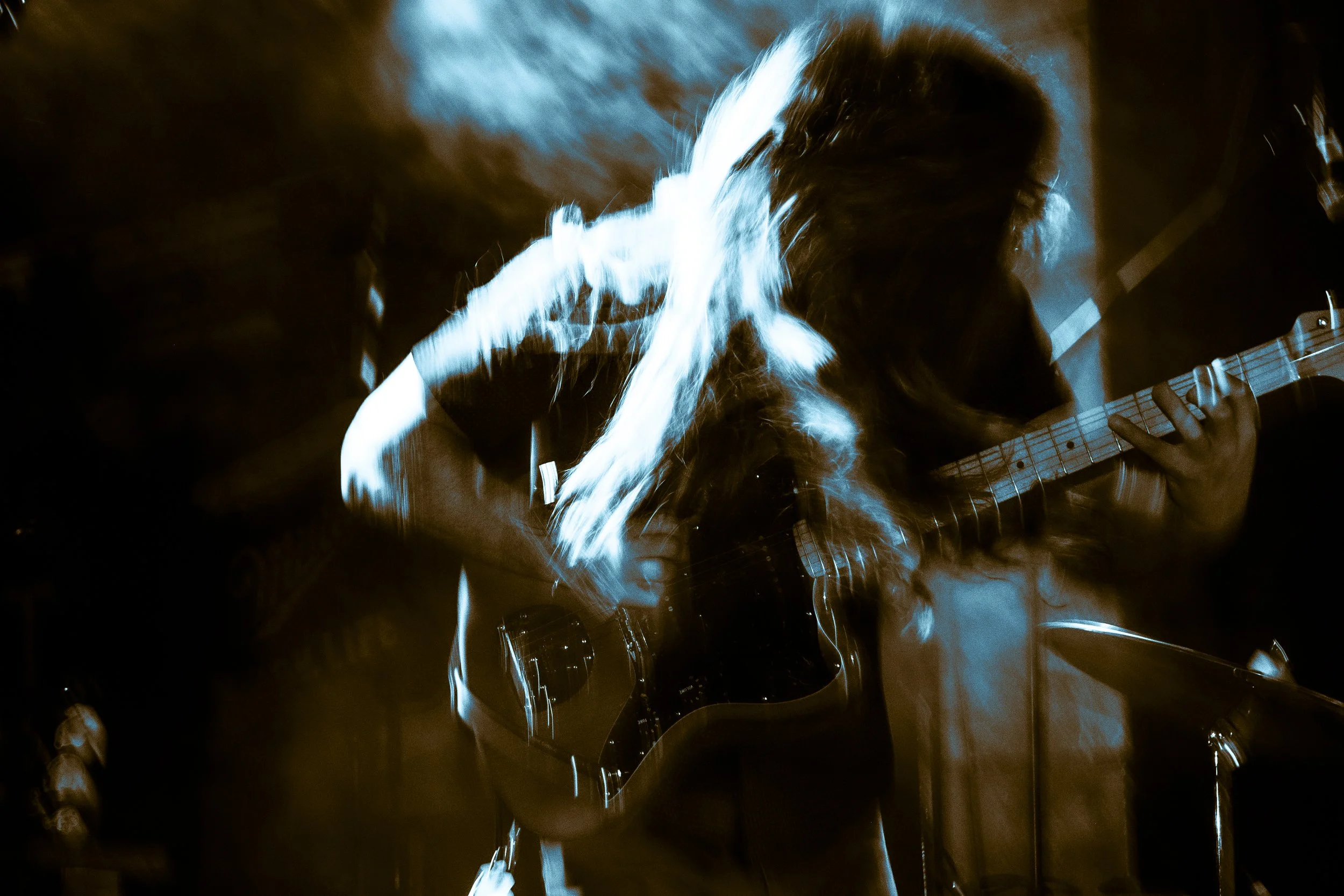 Liquid Pennies frontman/guitarist Chas BInns playing a Gibson SG electric guitar with long hair, illuminated by blue and orange stage lights, creating a motion blur effect.