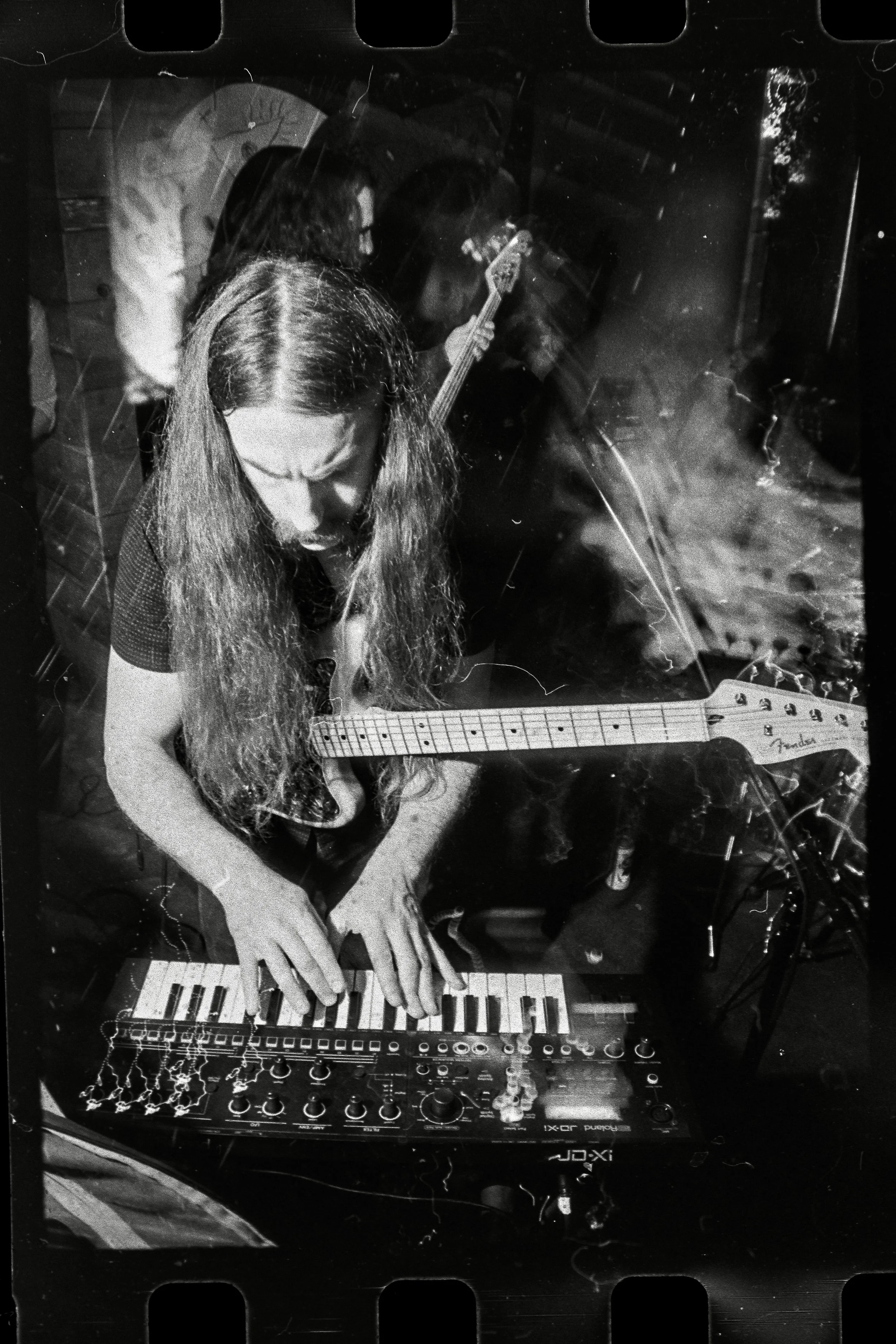Black and white photo of a woman playing a keyboard and a guitar during a live music performance, with another musician in the background.