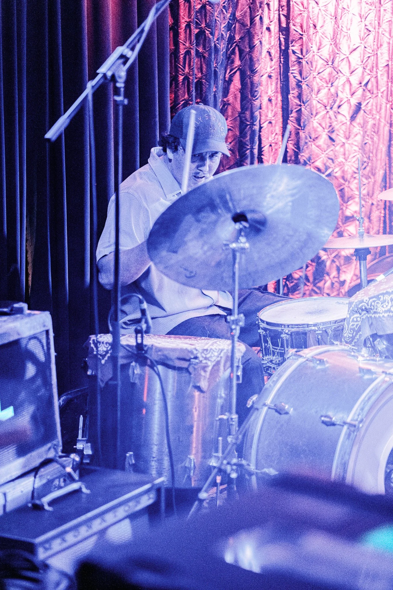 A man playing a drum set on stage with red and purple curtains in the background.