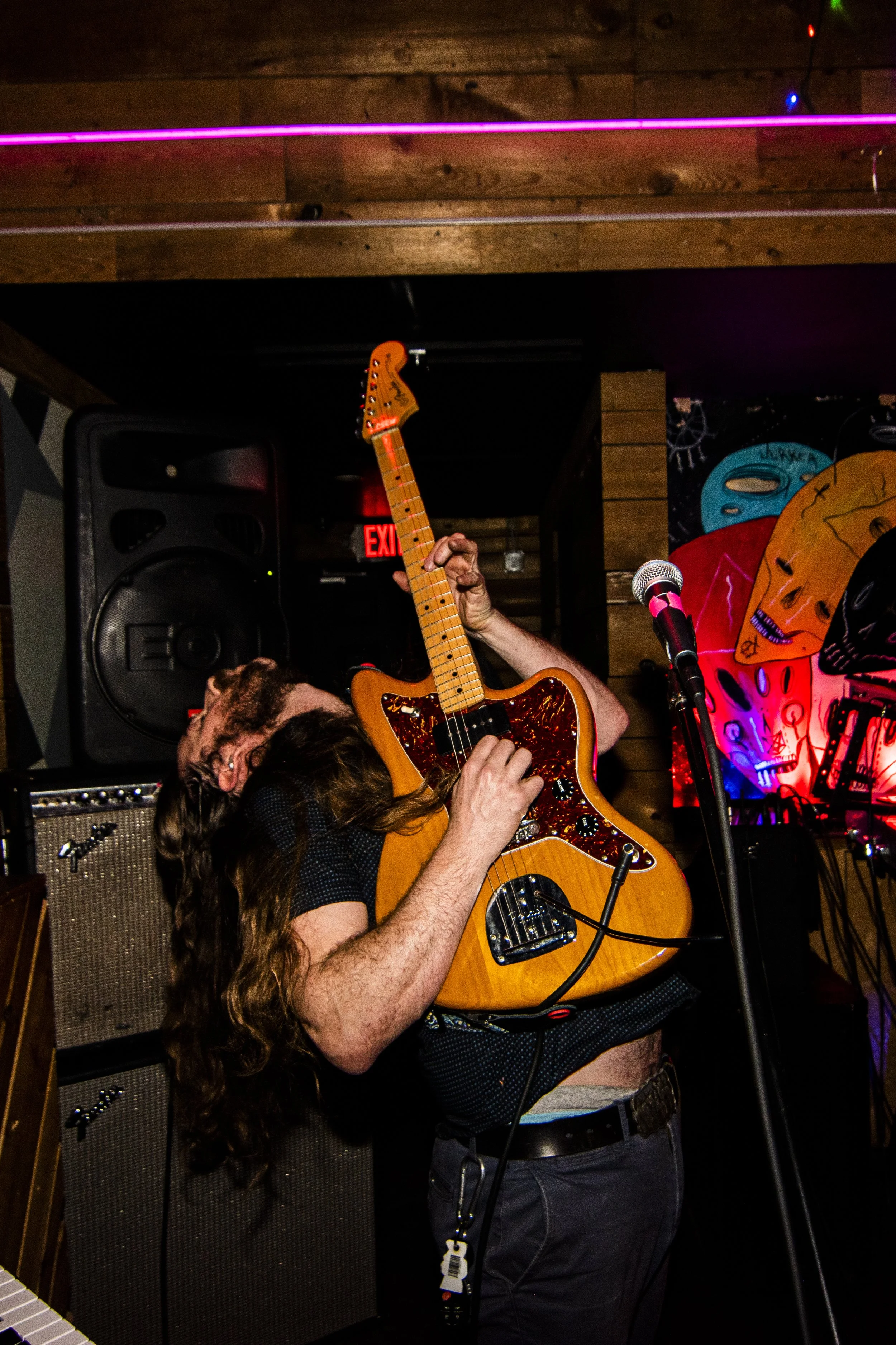 Man with long hair playing an electric bass guitar on stage in a dimly lit venue, with colorful artwork in the background.