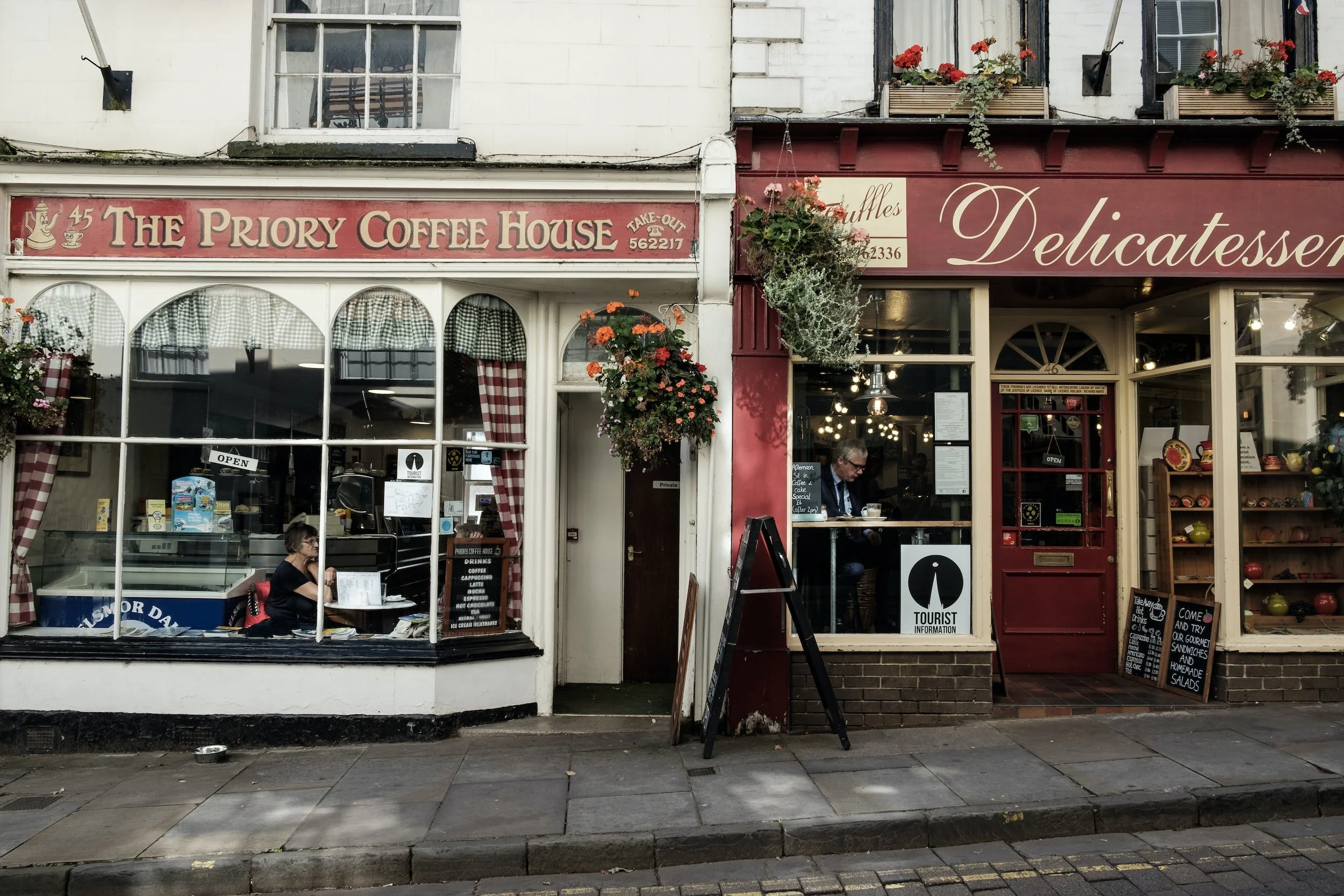 A street view showing two adjacent shops: The Priory Coffee House with a large front window and red sign, and a delicatessen with a burgundy facade and hanging flower baskets. Both shops have signs and outdoor chalkboards.