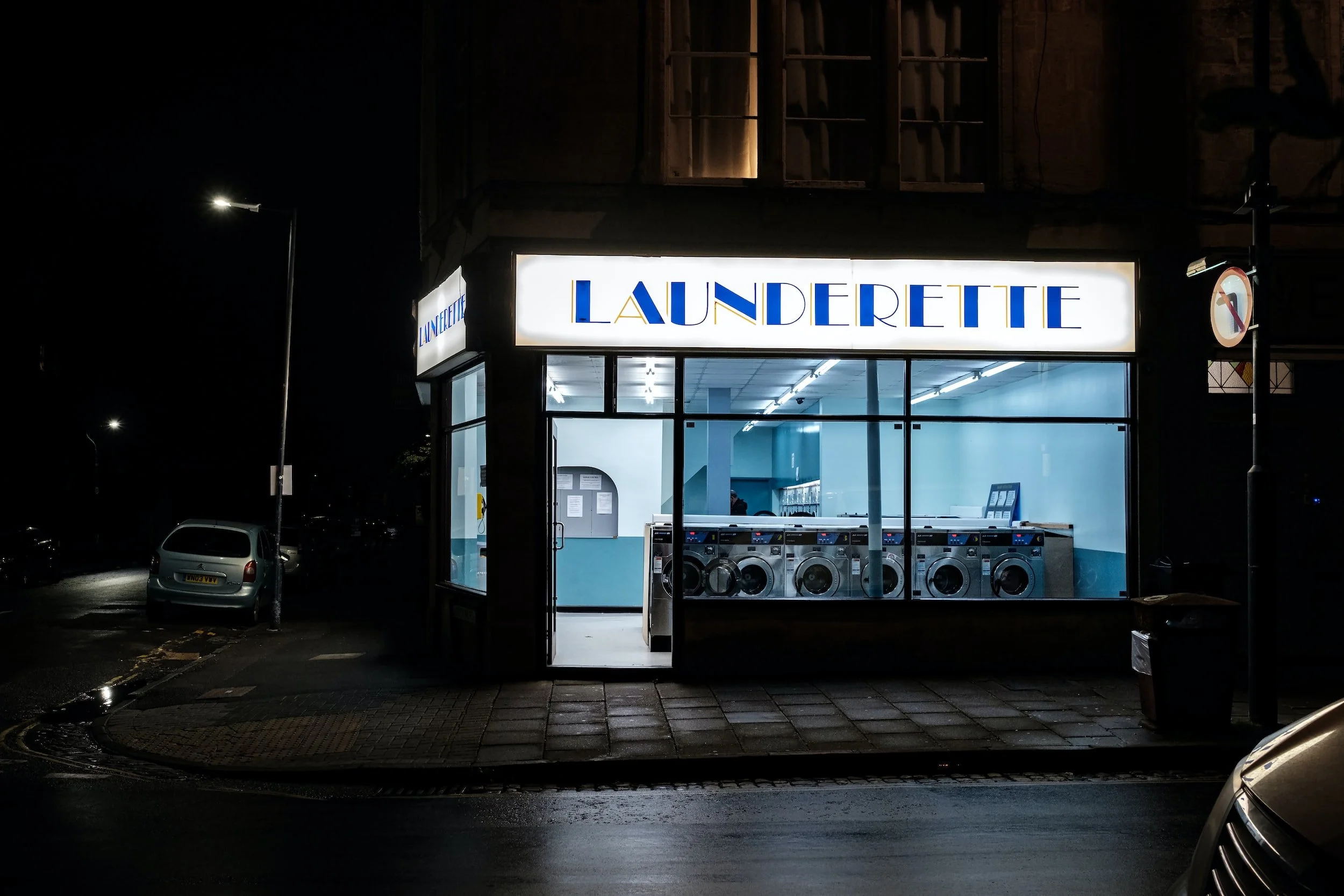 Night view of a laundromat named 'Launderette' with large illuminated sign, showing washing machines inside, illuminated blue walls, and parked cars outside.