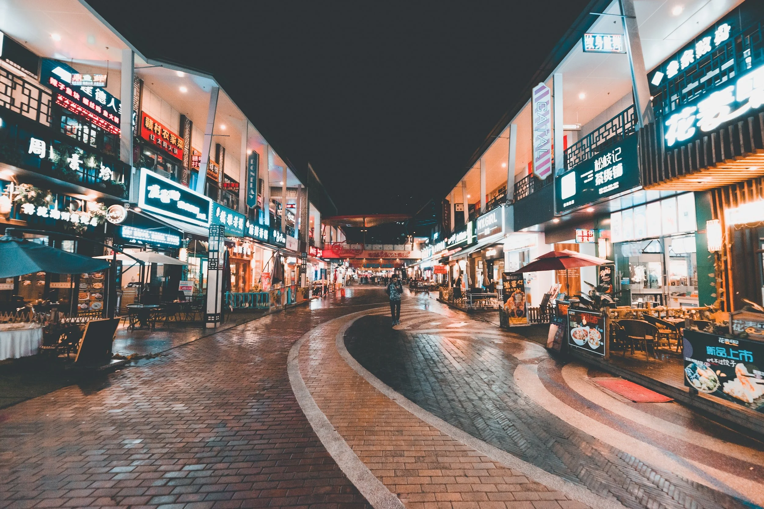 Empty street with shops and restaurants illuminated by neon signs at night, curved brick-paved walkway, no visible cars, one person walking in the center.