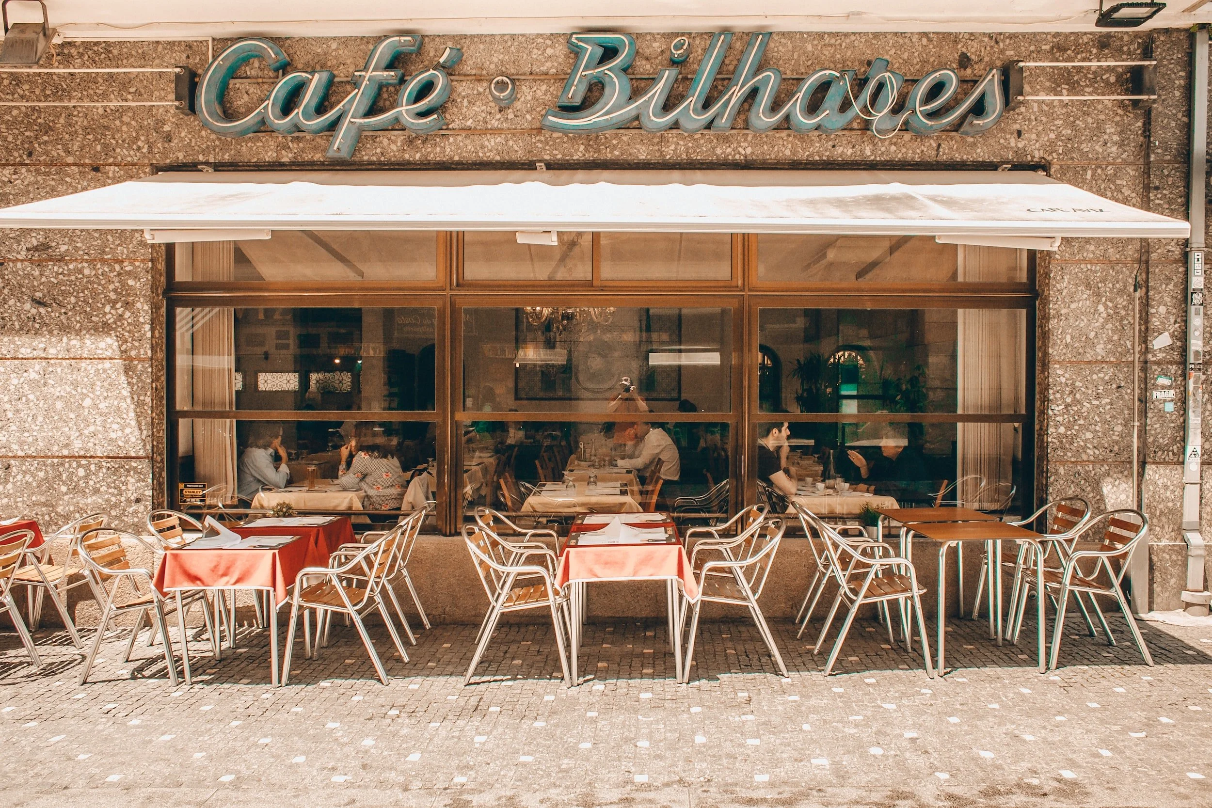 Exterior of a restaurant named Café Buharest with outdoor seating featuring several tables with red tablecloths and metal chairs. The large window shows people dining inside.