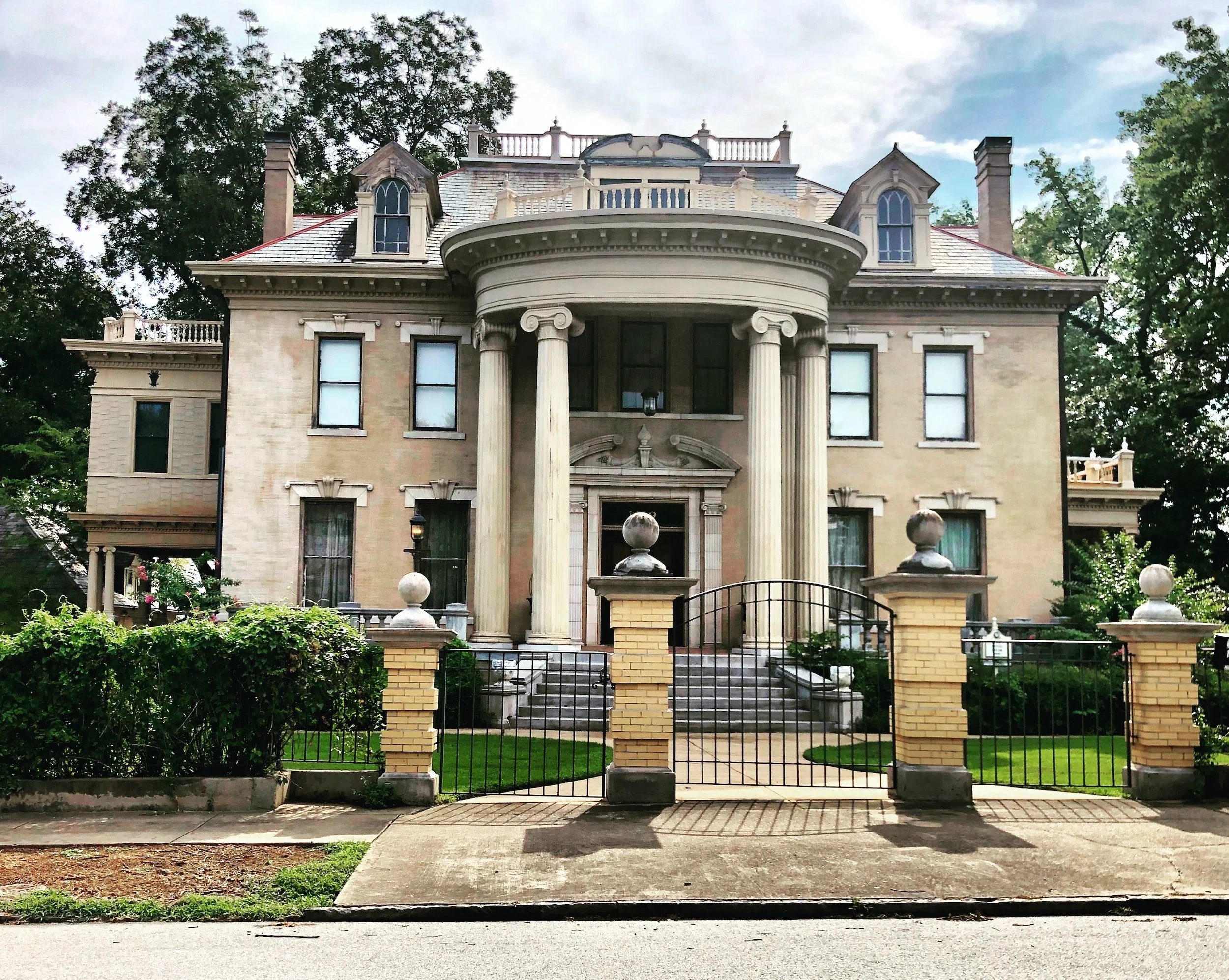 A large, historic mansion with a symmetrical facade, tall columns at the entrance, and a decorative staircase leading up to the front door. The house is surrounded by a black wrought iron gate and a small front yard with green grass and bushes.