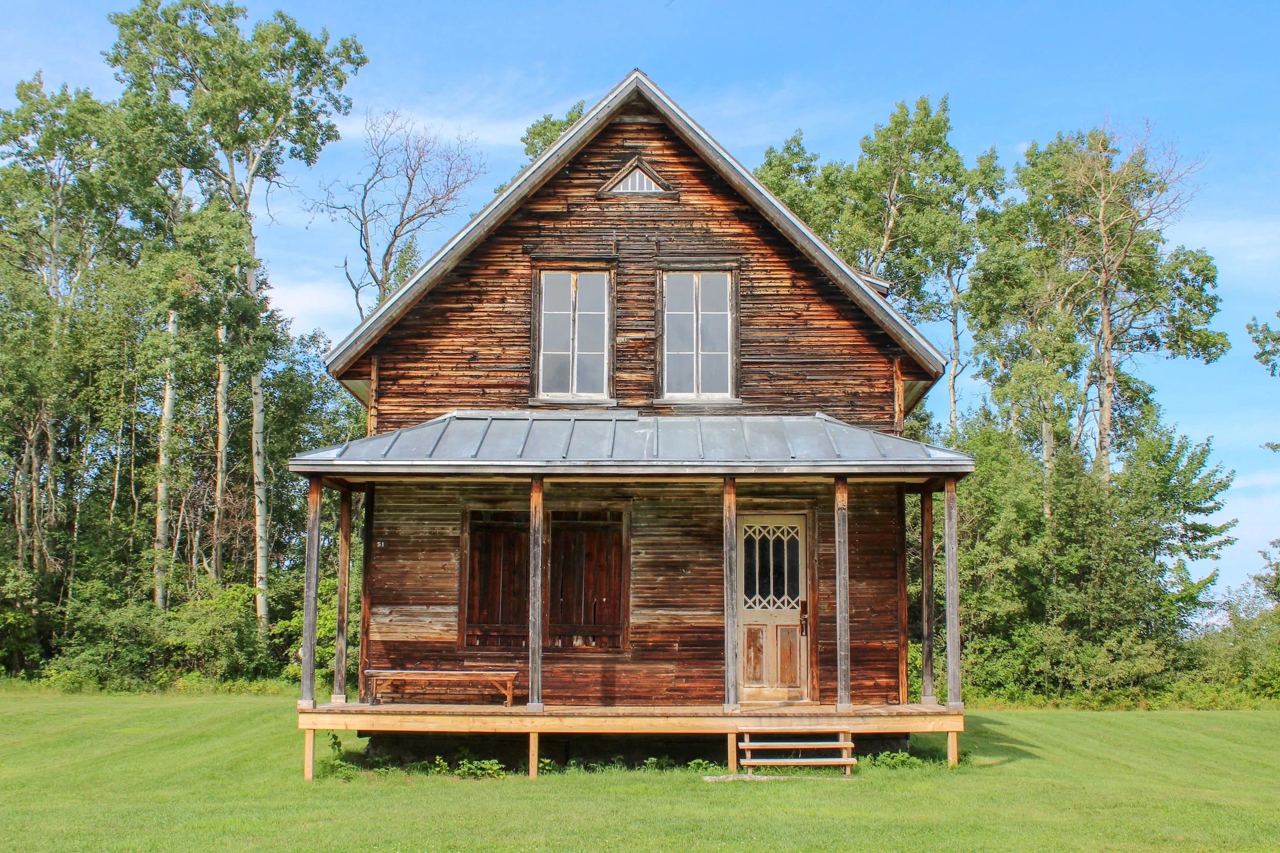 Old wooden house with a porch, surrounded by green grass and trees, under a blue sky with some clouds.