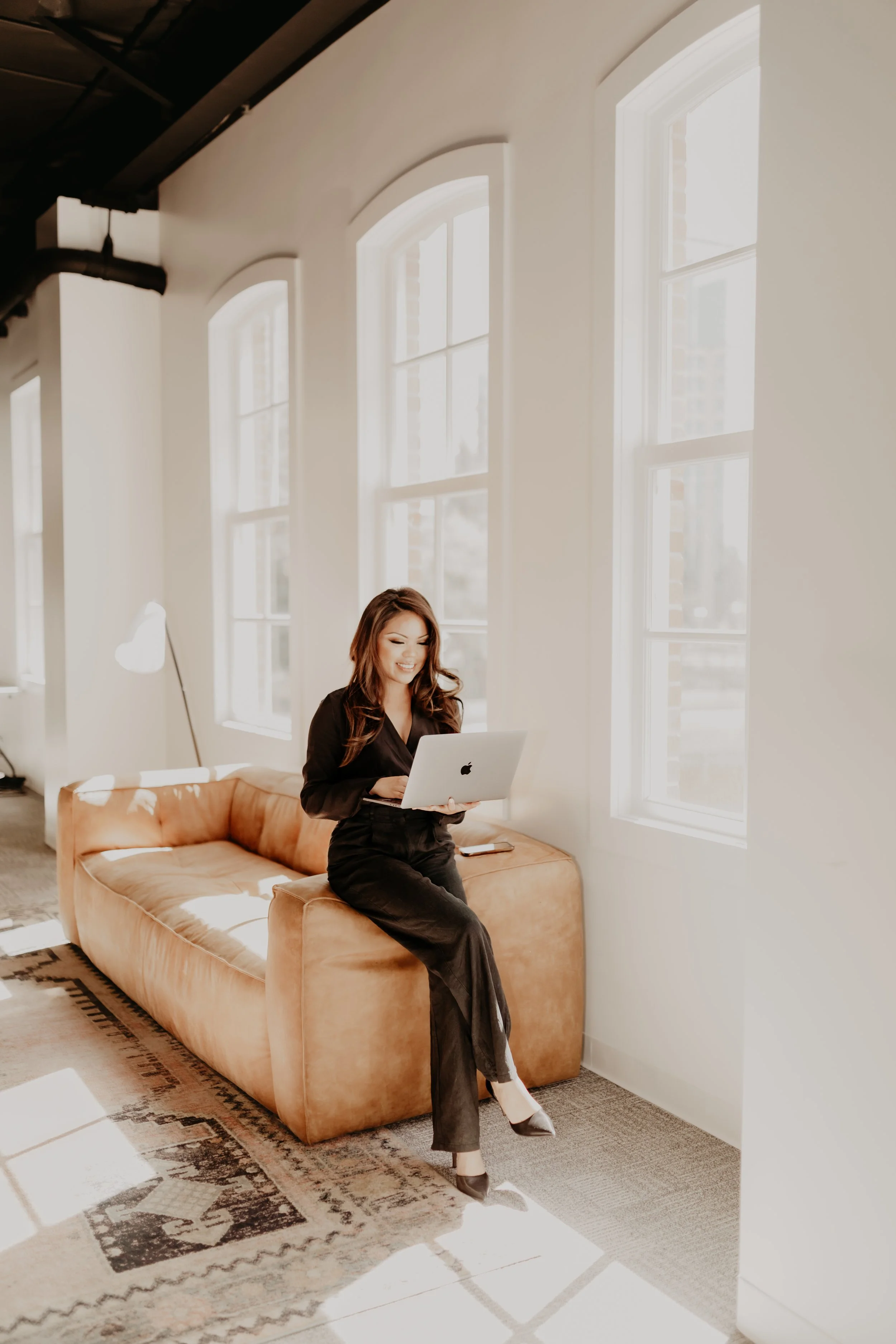 A woman sitting on a tan velvet sofa, working on a silver Apple MacBook laptop, in a brightly lit room with large arched windows and a patterned rug.