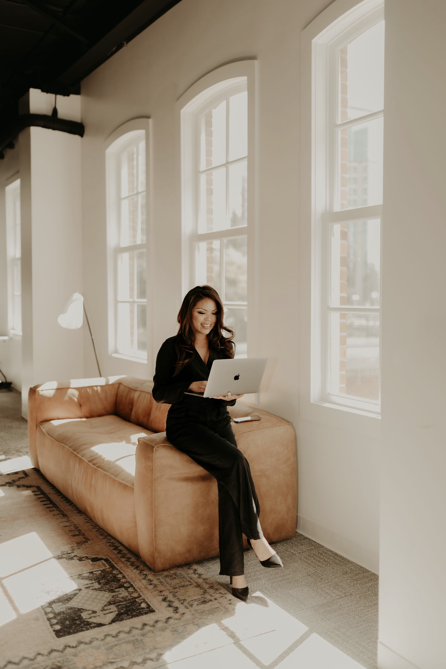 A woman in black clothing sitting on a tan couch with a laptop, in a room with large windows allowing sunlight, a patterned rug, and a white wall.