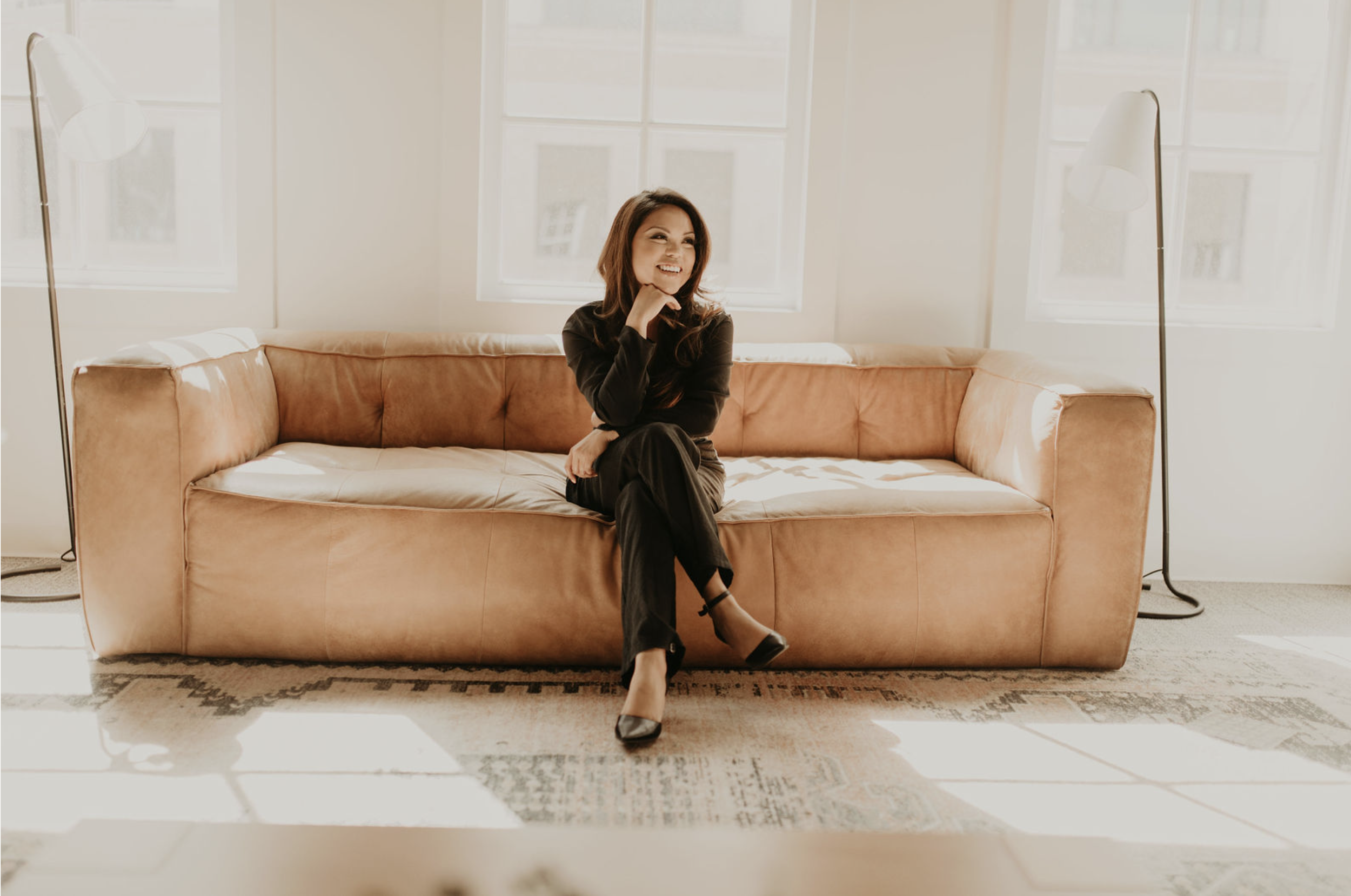 Woman sitting on a beige sofa, smiling, wearing black clothing and heels, in a bright room with large windows and two tall floor lamps.